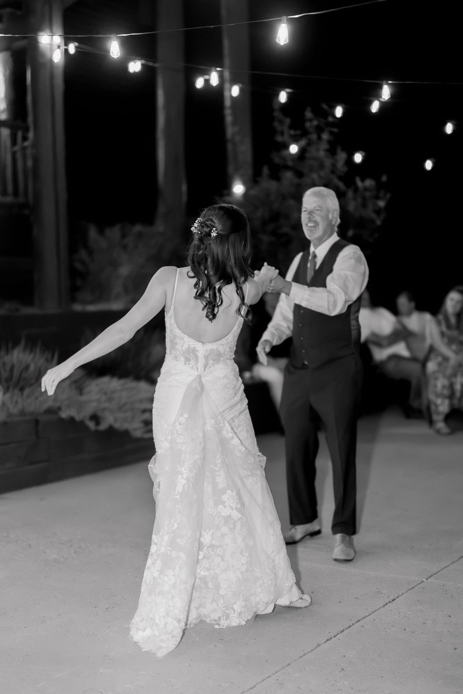 Black and white photo of a bride in lace gown dancing with an older man under string lights at an outdoor wedding reception.