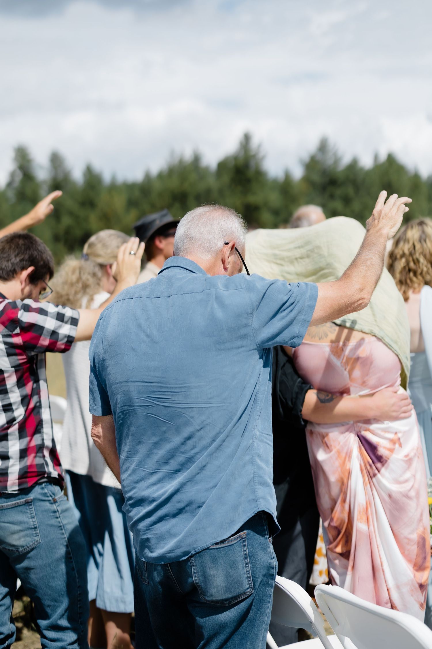 Outdoor wedding guests raising hands in celebration, surrounded by evergreen trees under a cloudy sky.