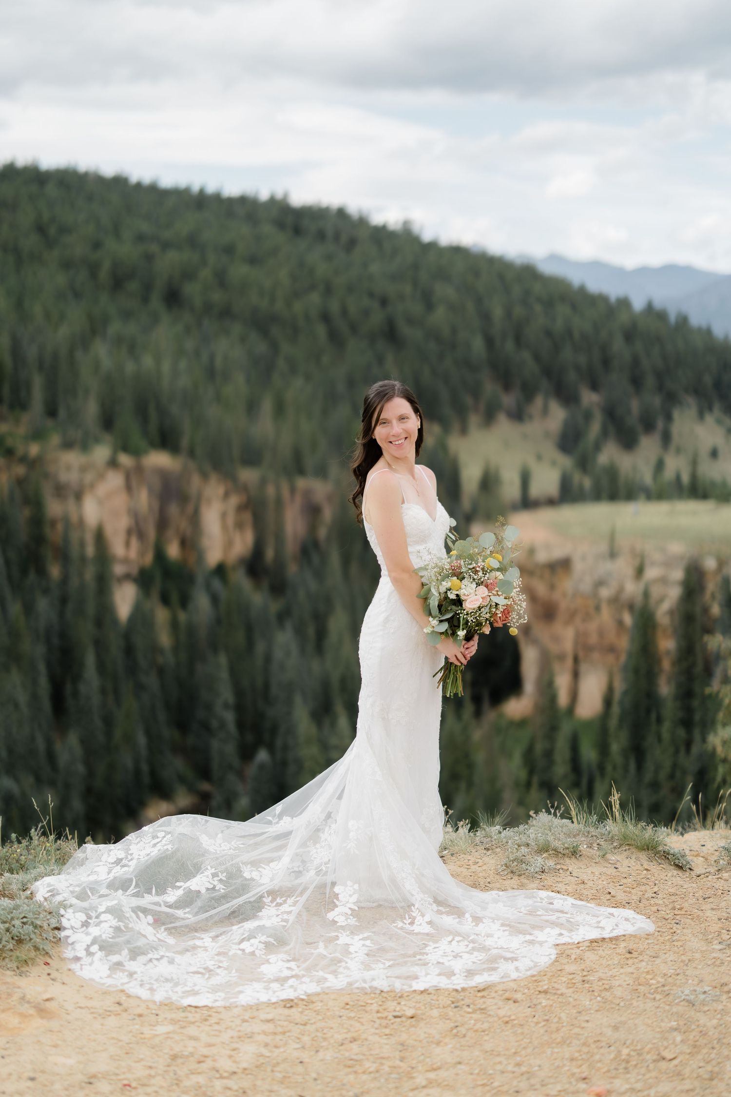 Bride in lace gown with train holds bouquet on mountain overlook with pine forest backdrop.