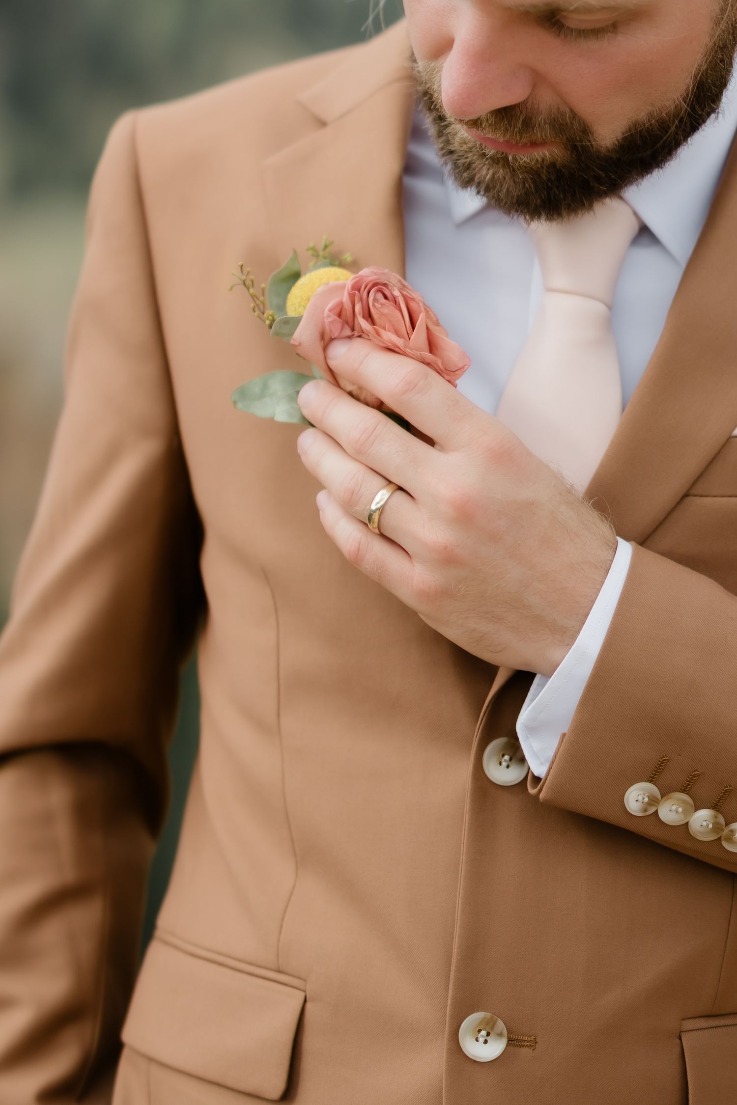 Groom in tan suit adjusting pink rose boutonniere, wearing gold wedding band, white shirt and blush tie.