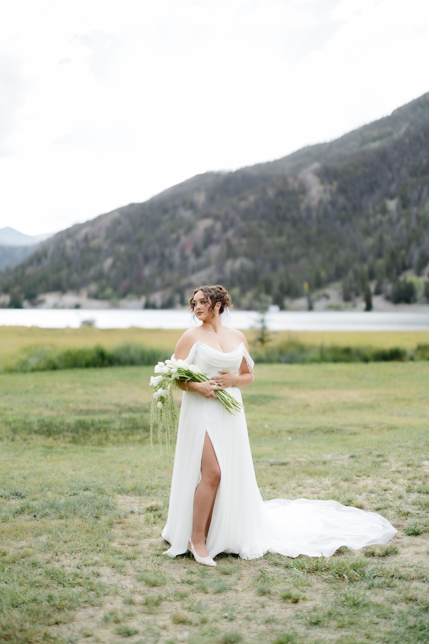 Bride in white gown with slit holds bouquet in mountain meadow near lake.