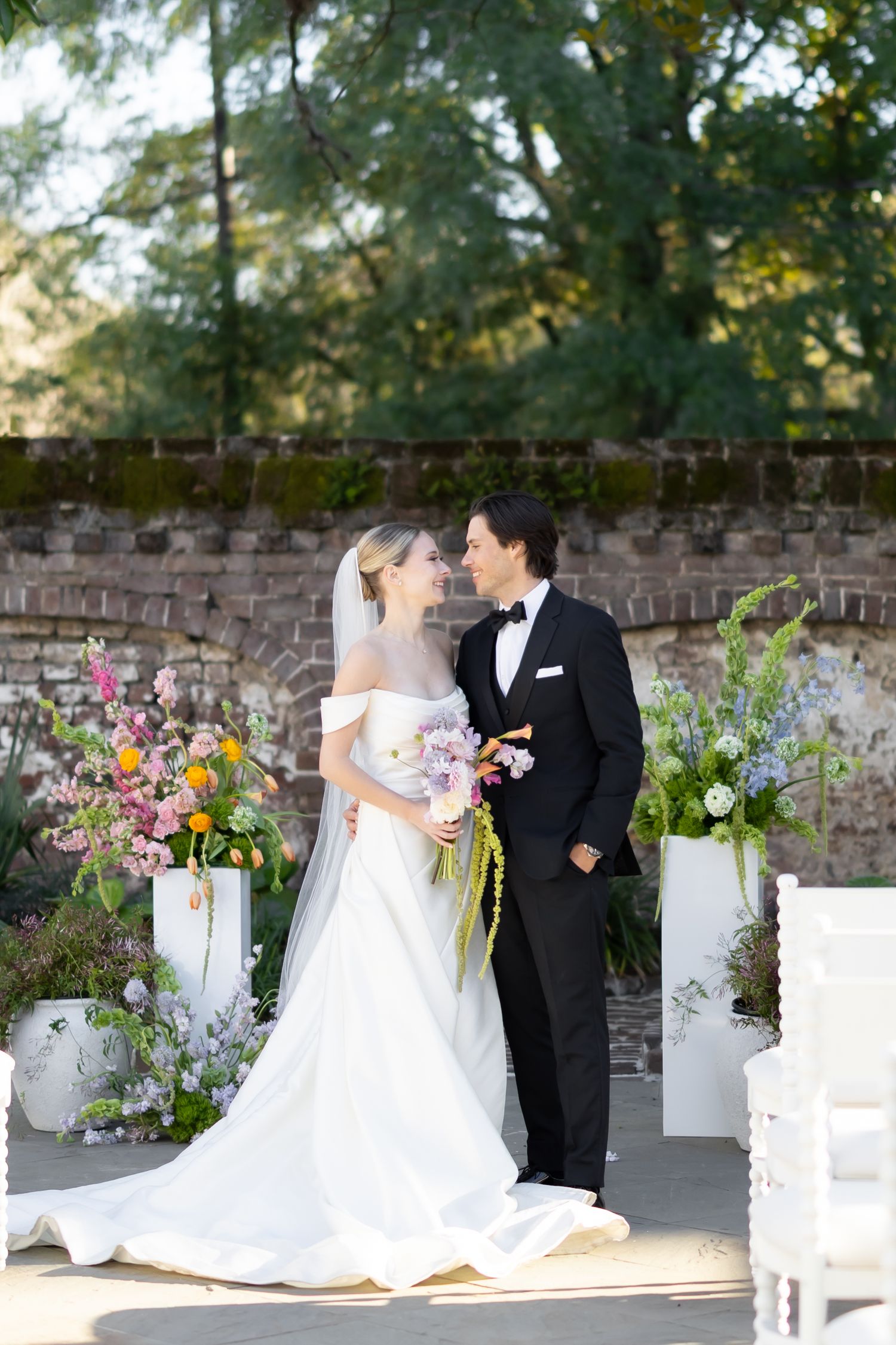 Wedding couple in elegant attire stand together at outdoor ceremony with colorful floral arrangements.