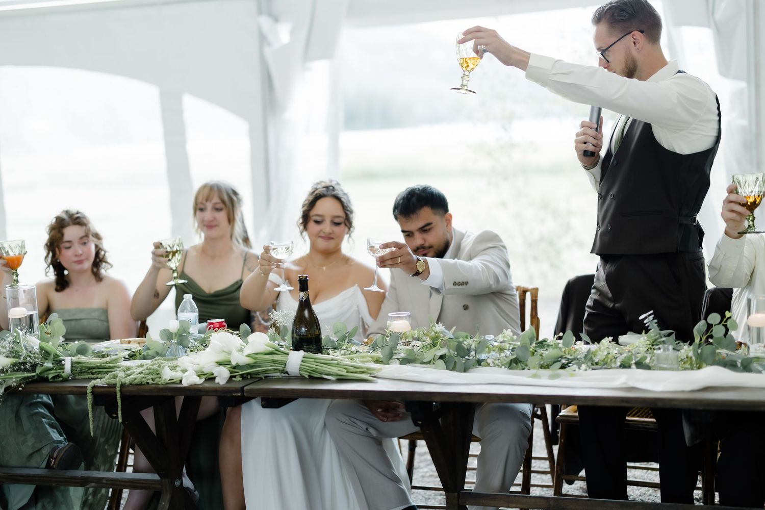 Wedding party raises champagne glasses in a toast at a decorated reception table with greenery and floral arrangements.