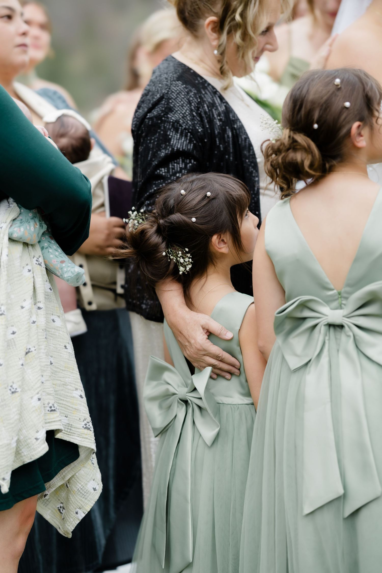 Flower girls in sage green dresses with large bows stand together at outdoor wedding ceremony.