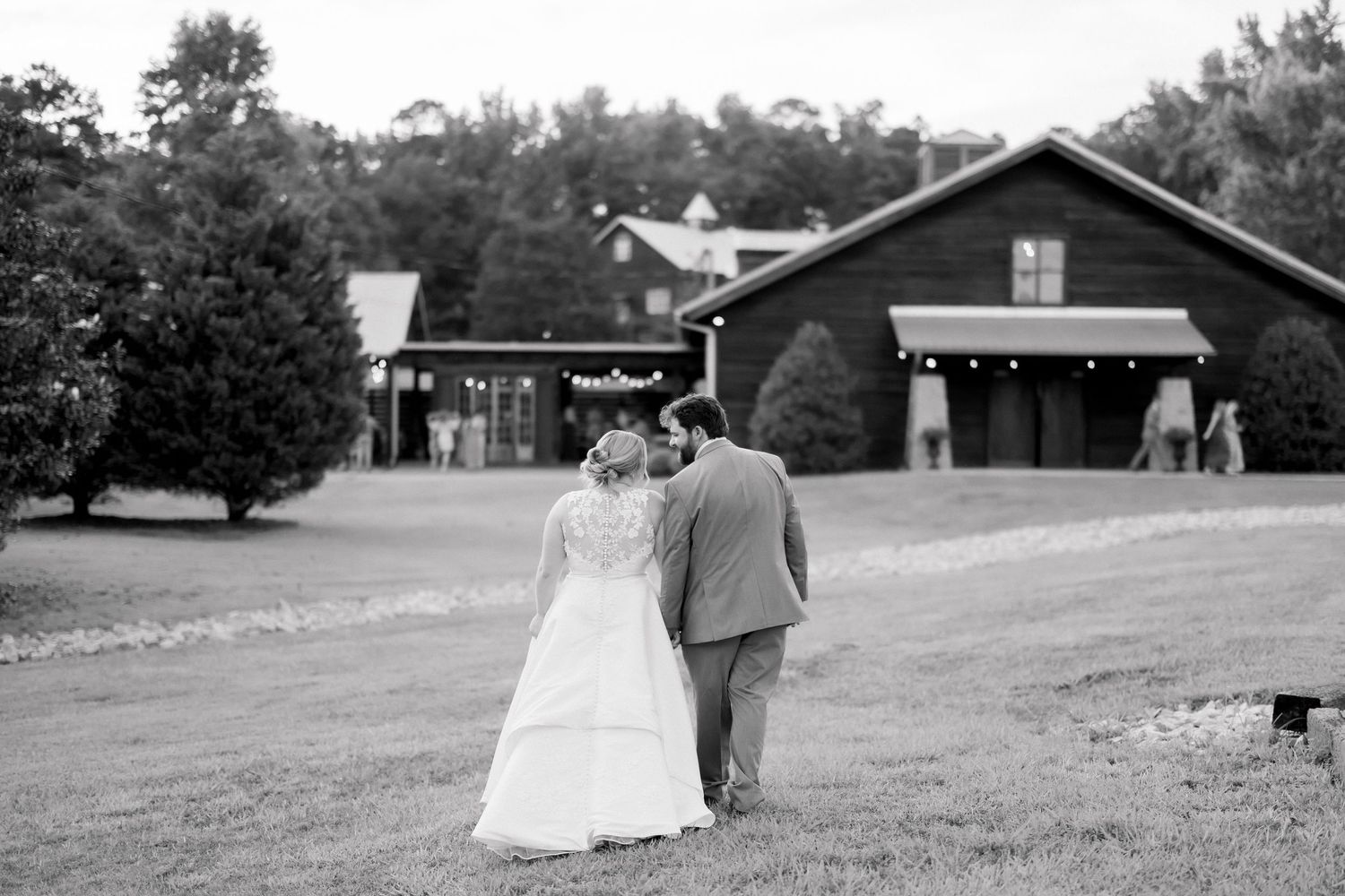 Black and white photo of a bride and groom walking toward a rustic barn venue on their wedding day.