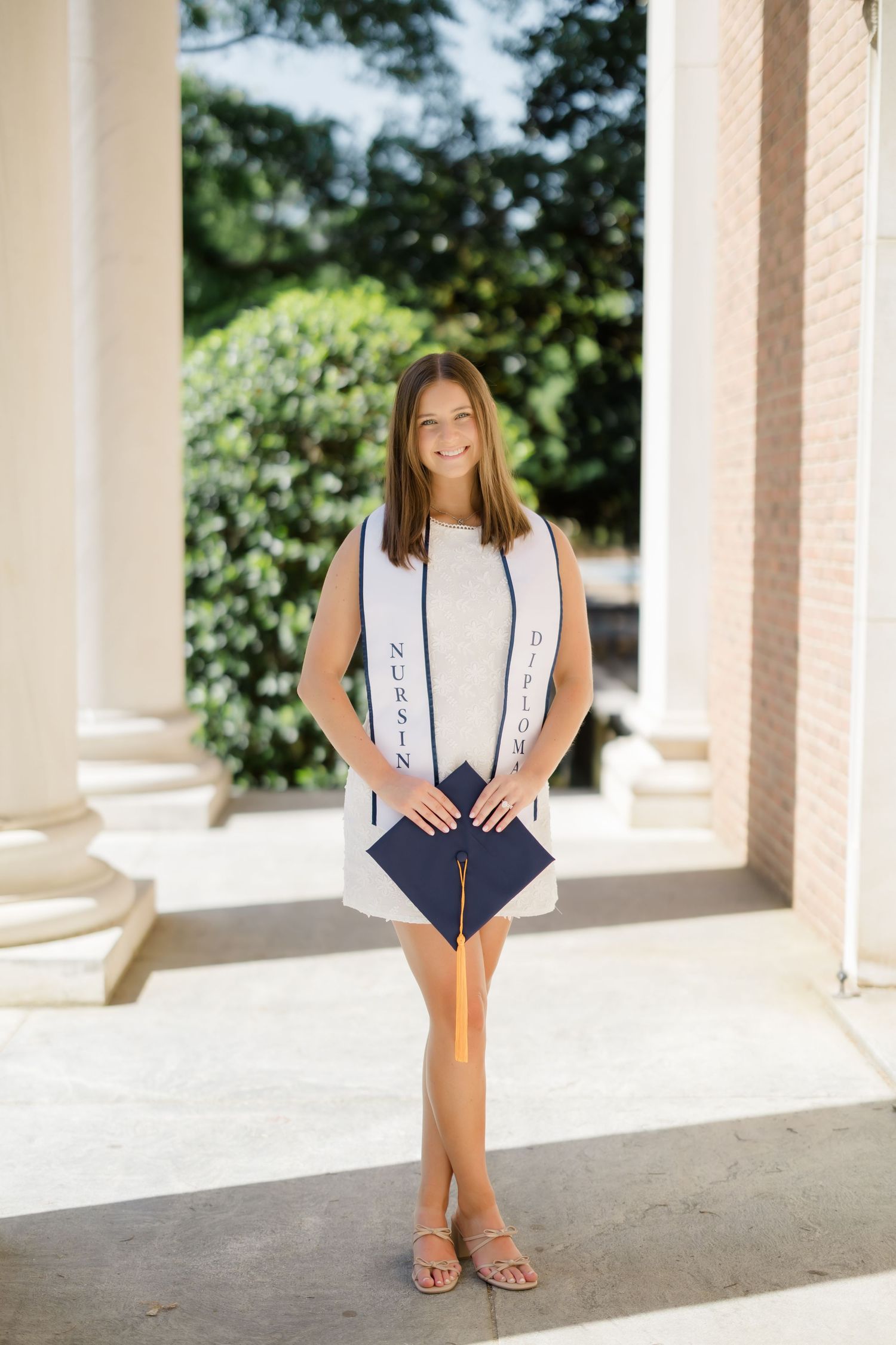 Young woman in white nursing graduation stole holds navy cap, posing between columns outdoors.