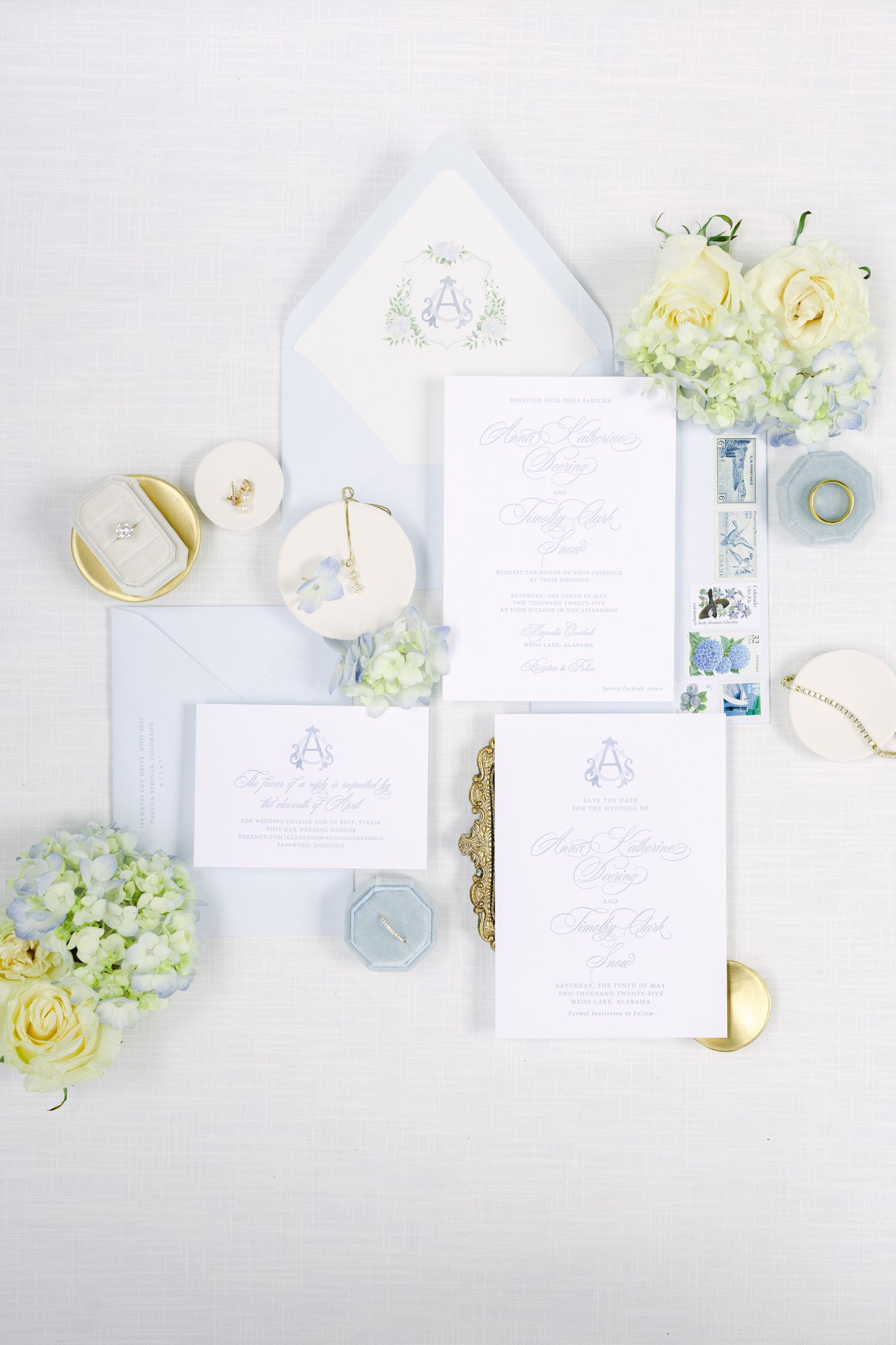 Wedding stationery flatlay with elegant white and blue invitations, pale green hydrangeas, and cream roses on light background.
