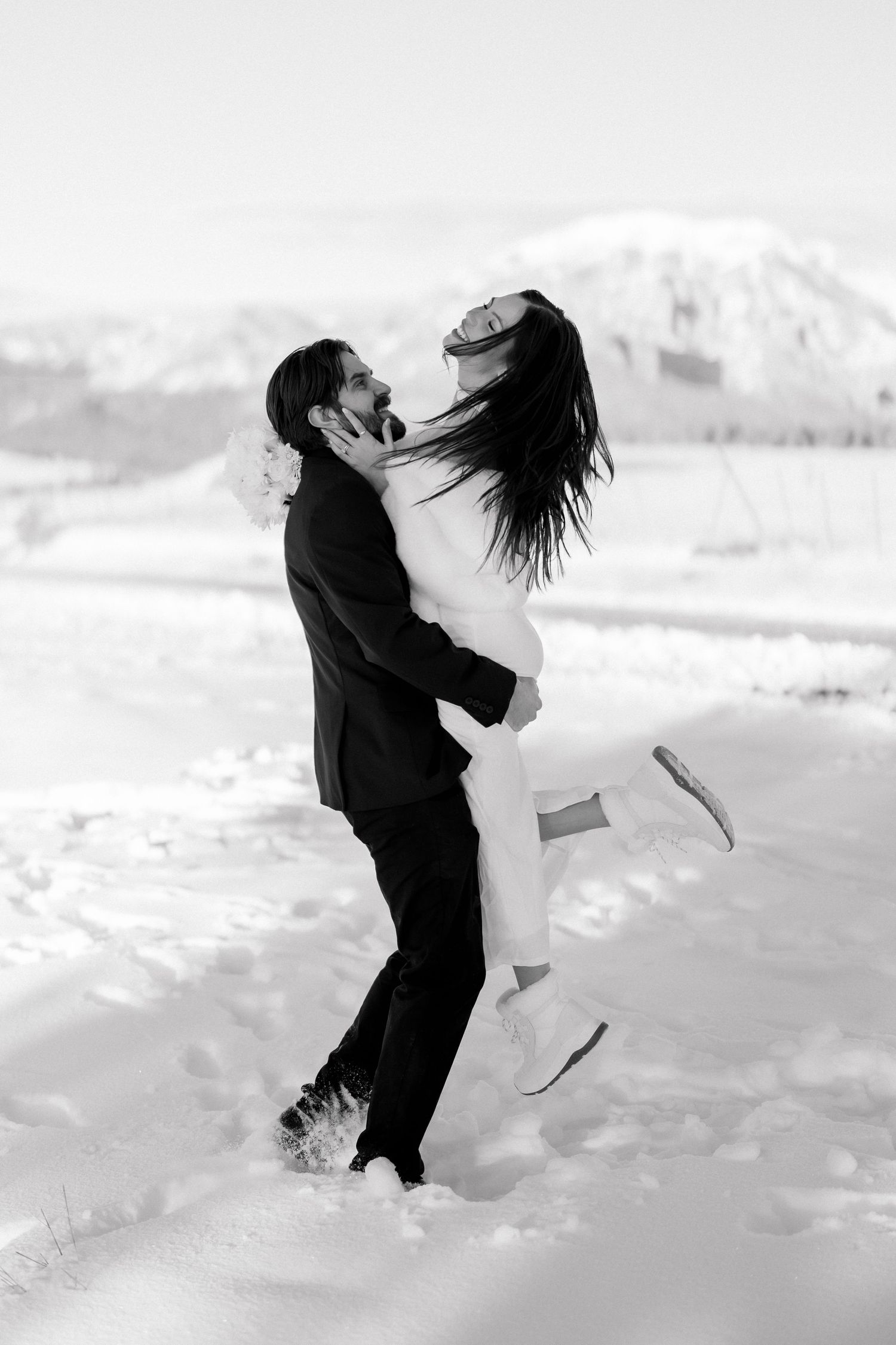 Groom lifts laughing bride in white fur stole in a joyful black and white winter wedding photo in the snow.