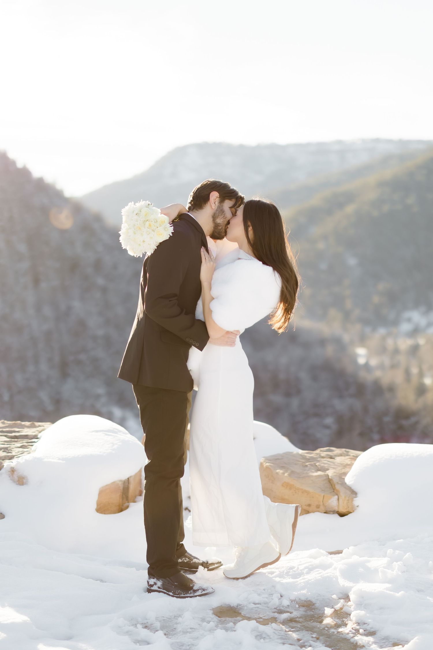 Bride and groom share a kiss in a snowy mountain landscape, bride holding white bouquet in winter wedding attire.