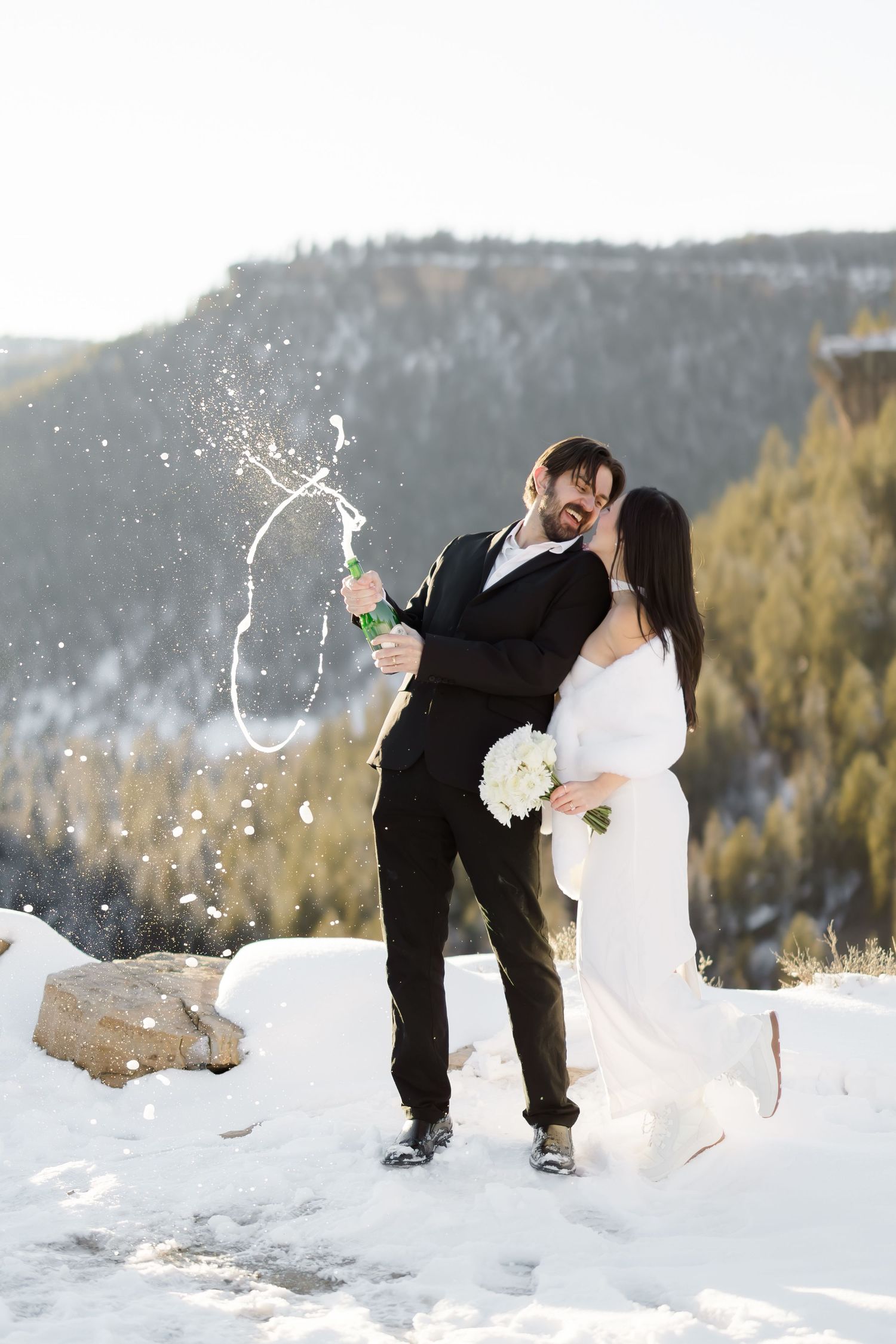Newlyweds celebrate by popping champagne bottle in snowy mountain setting with pine trees.