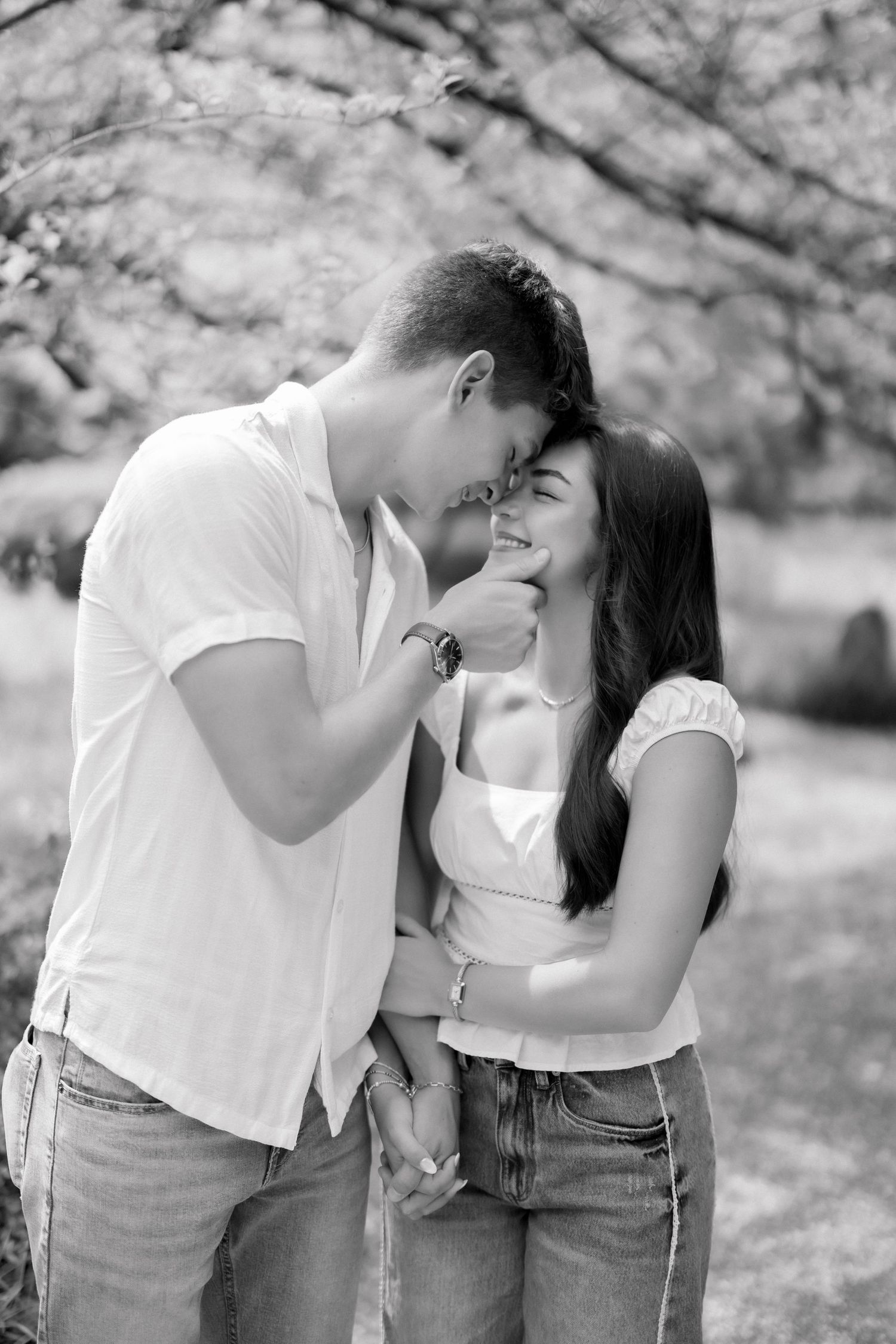 Black and white photo of a young couple sharing a tender moment, smiling and holding hands outdoors.
