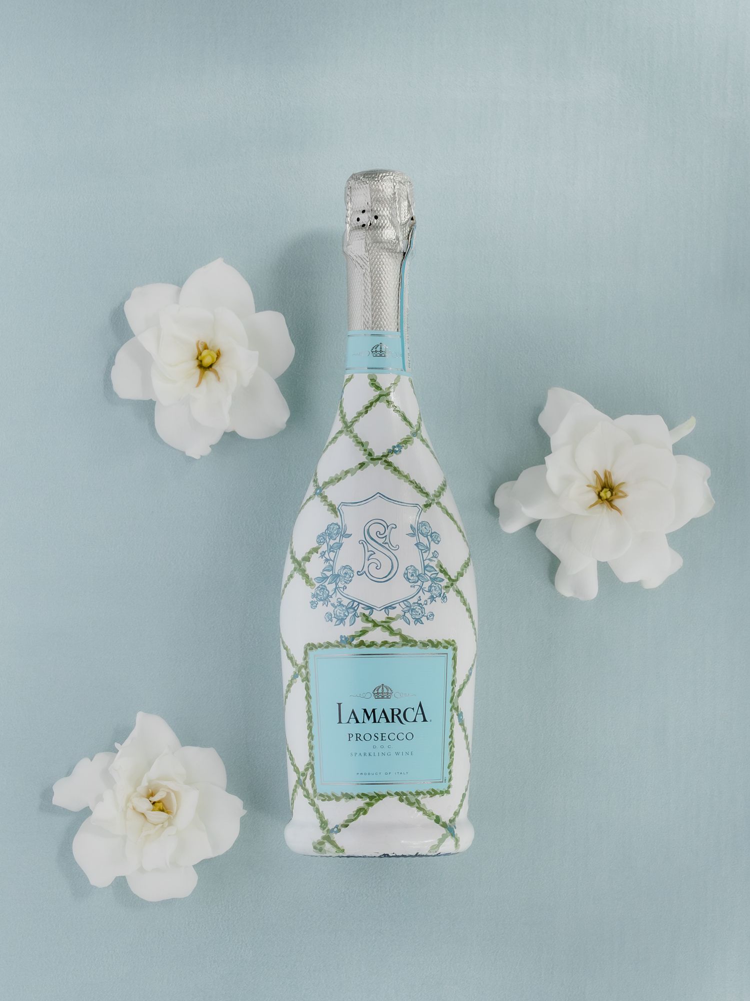 La Marca Prosecco bottle with floral embroidery design surrounded by white flowers on a soft blue background.