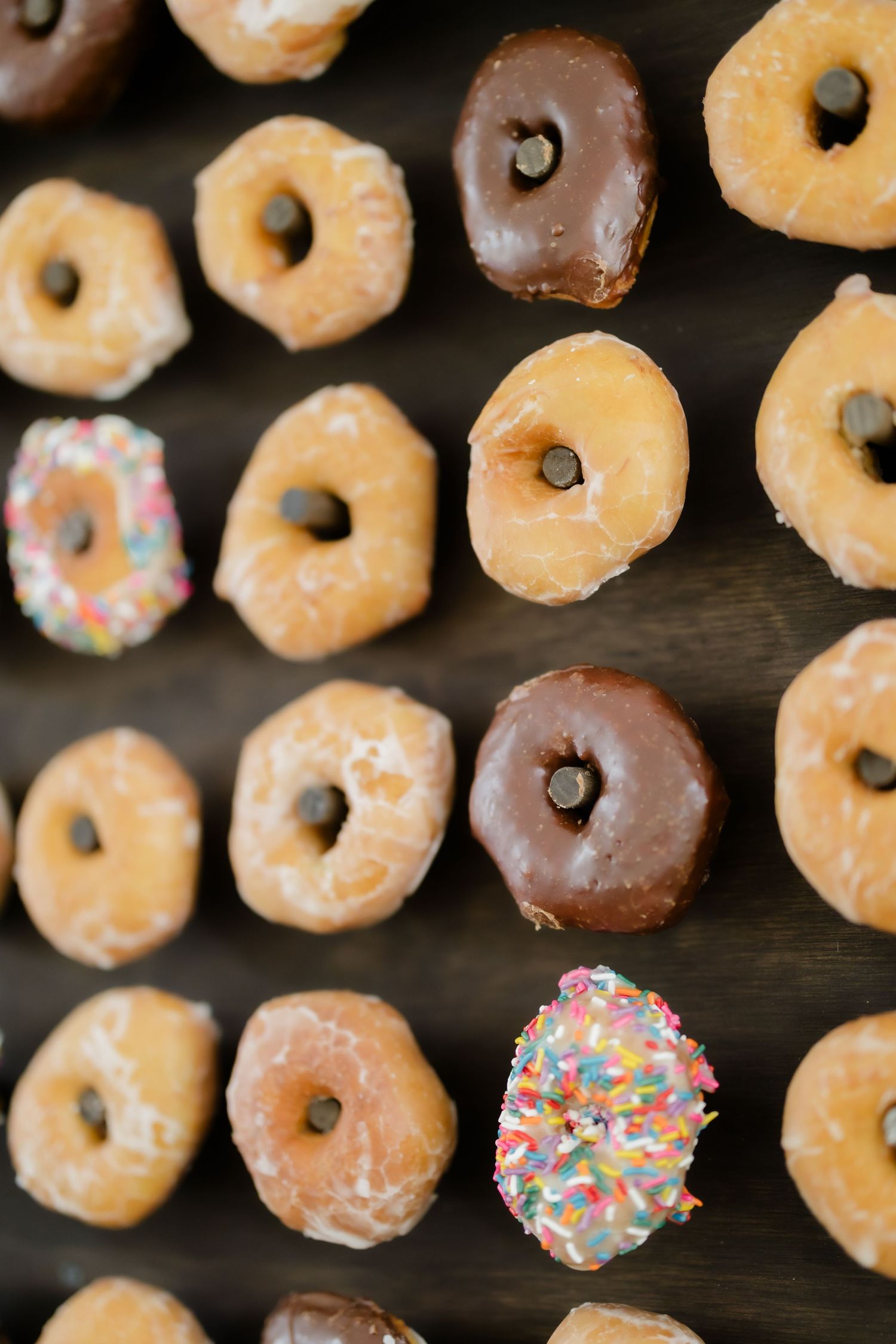 A display of assorted donuts including glazed, chocolate, and sprinkled varieties arranged in rows on a dark wooden surface.