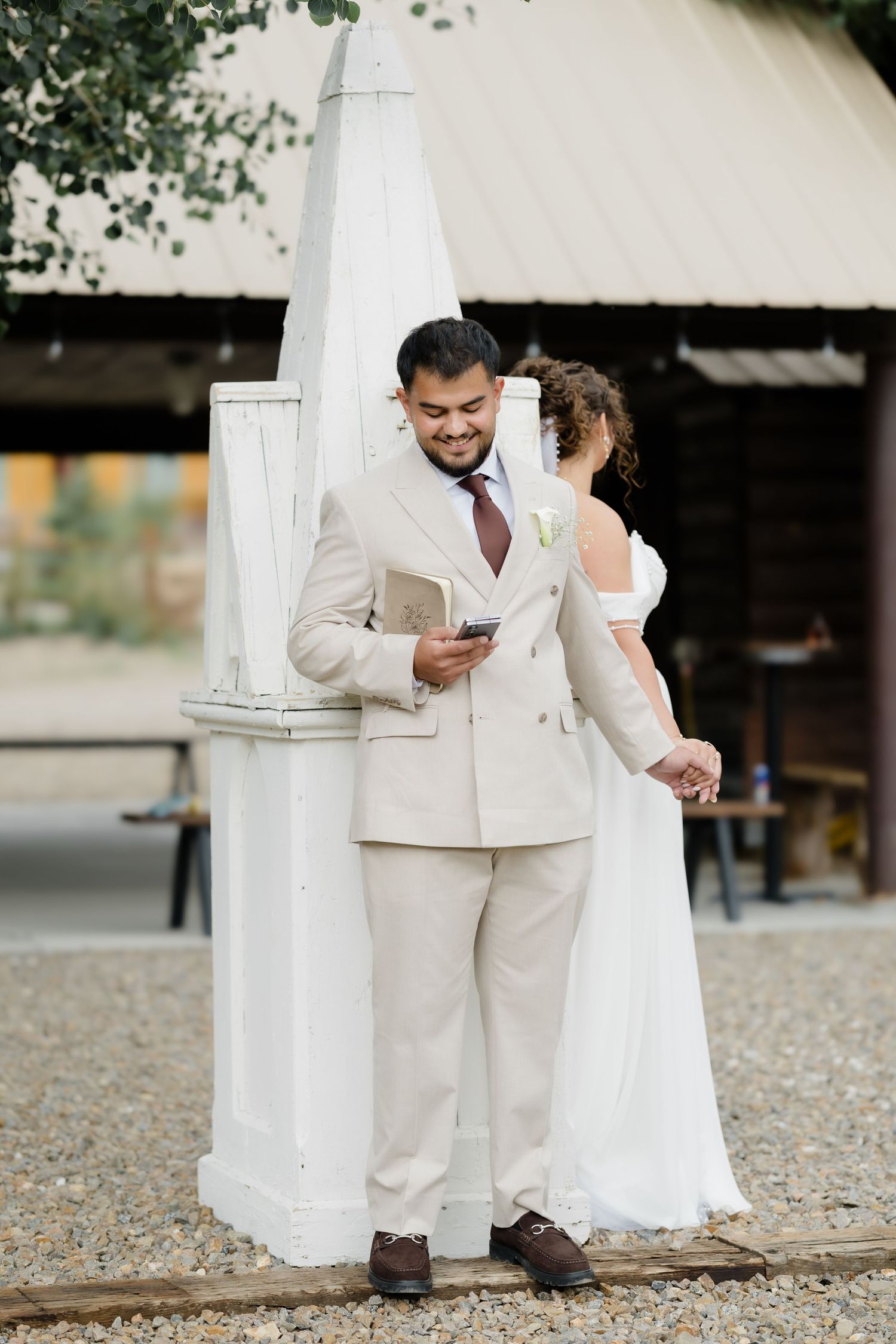 Groom in beige suit checks phone while bride in white dress holds his hand at outdoor wedding venue.