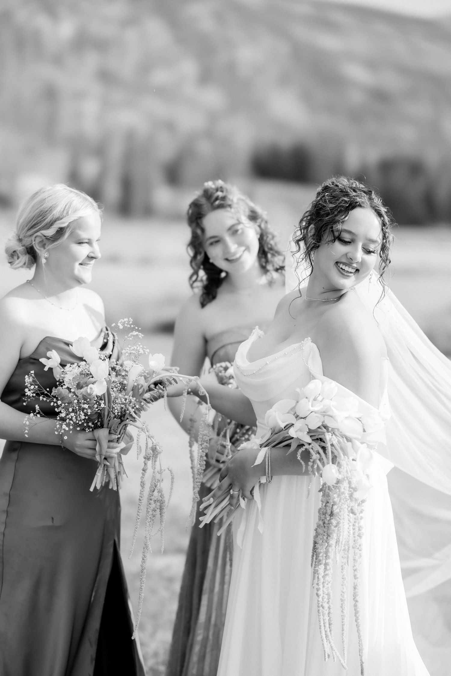 Black and white photo of bride in white gown smiling with two bridesmaids holding bouquets outdoors.