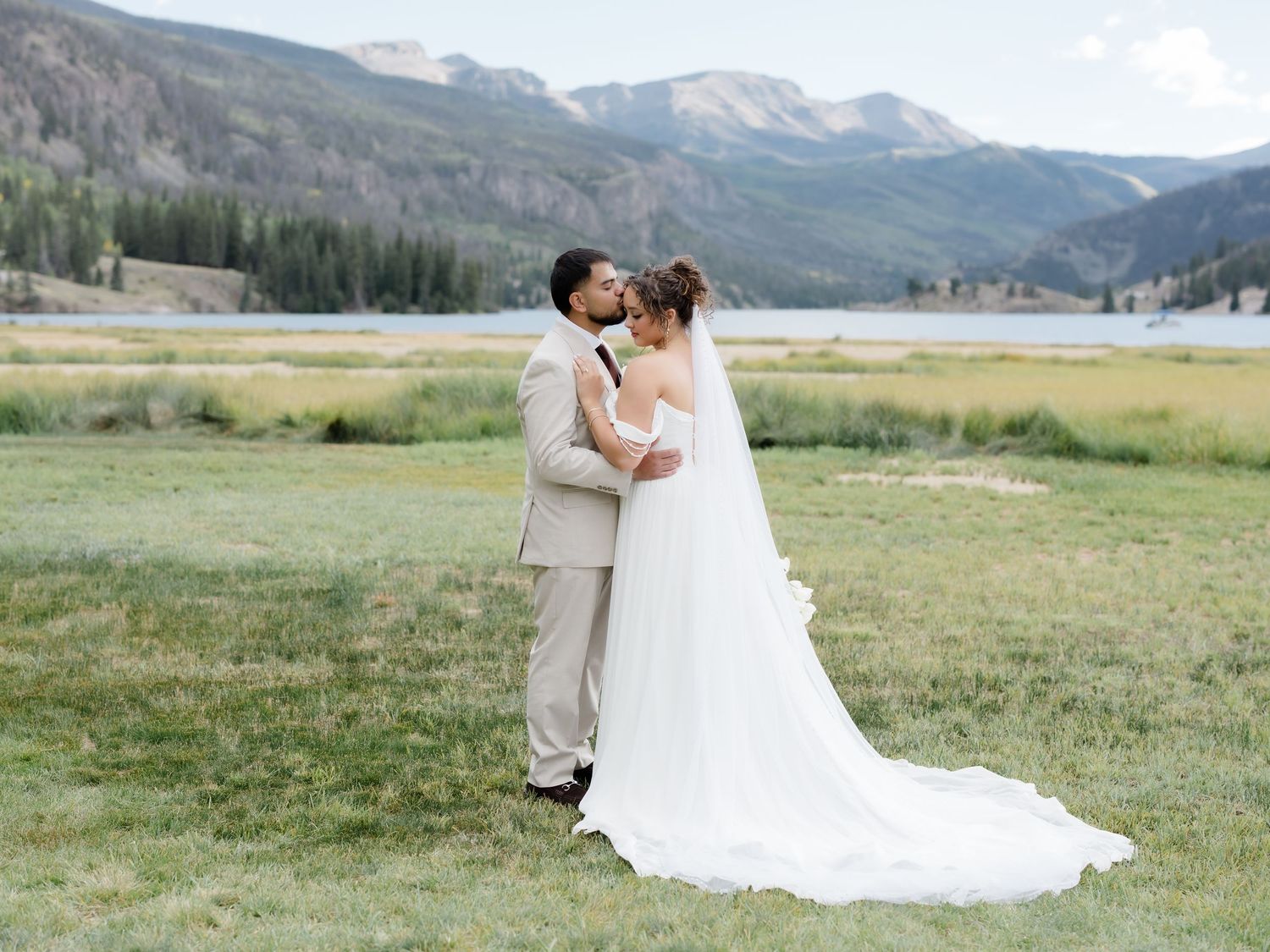A bride and groom share a kiss in a scenic mountain meadow, with a lake and pine trees in the background.