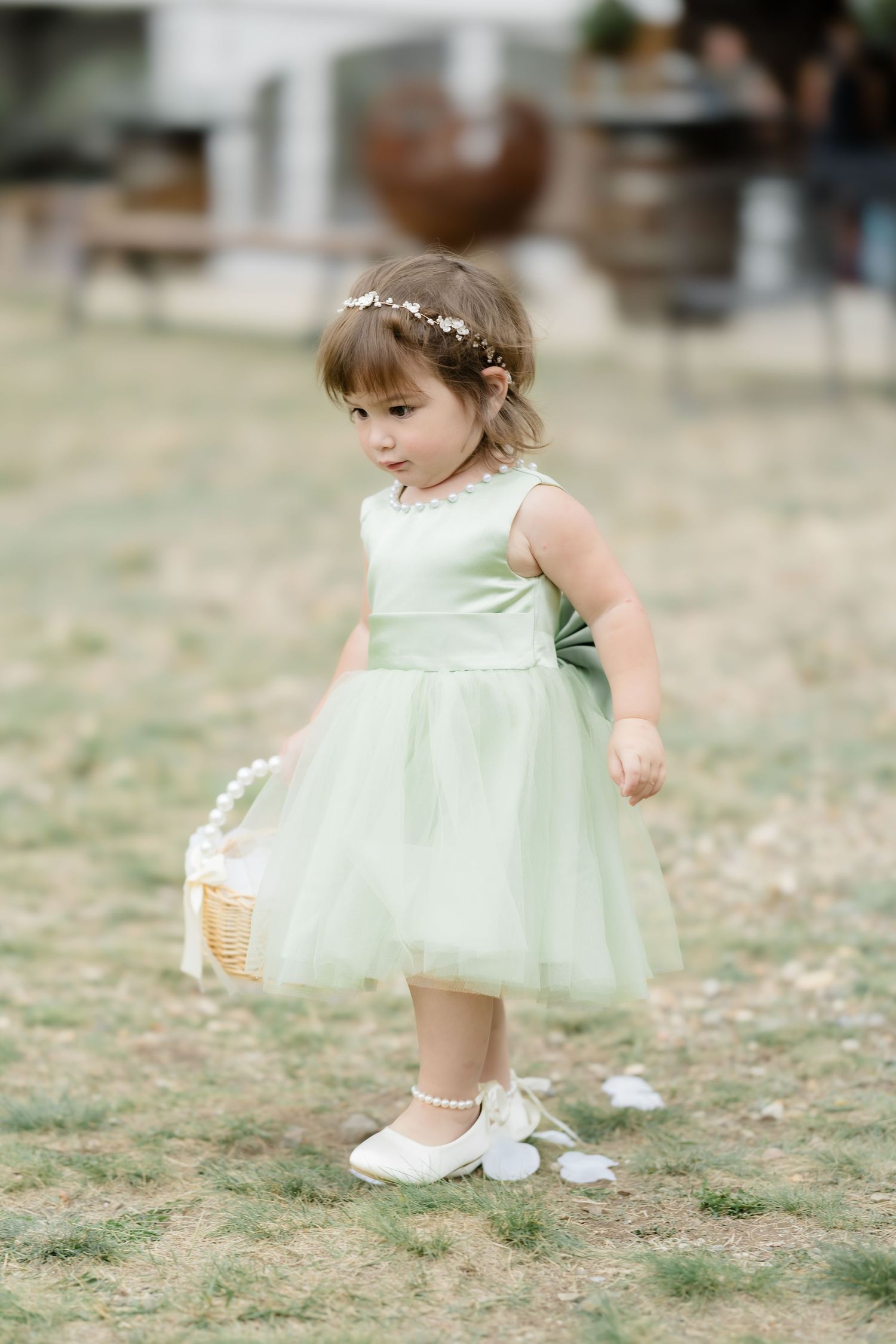 Adorable flower girl in mint green tulle dress with floral crown holding a basket at an outdoor wedding ceremony.