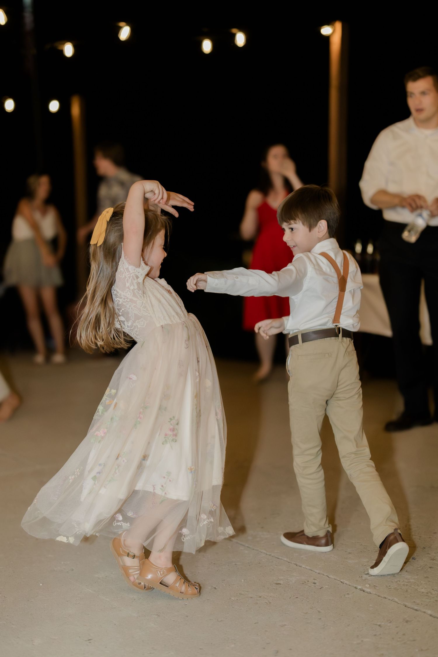 Two children dance together at a wedding reception, girl in floral dress and boy in suspenders.