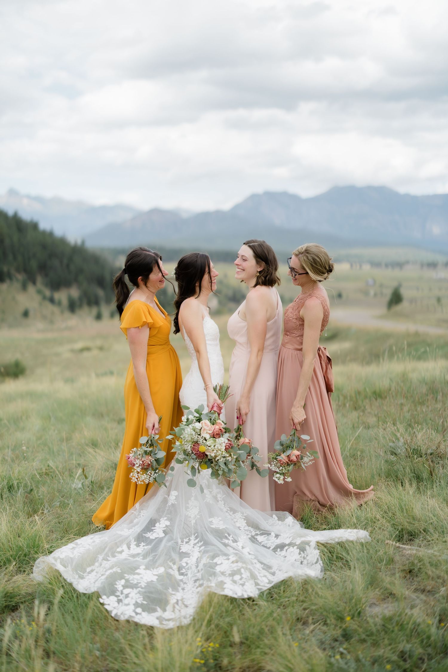 Bride in lace gown with bridesmaids in mustard and blush dresses holding bouquets in a mountain meadow.