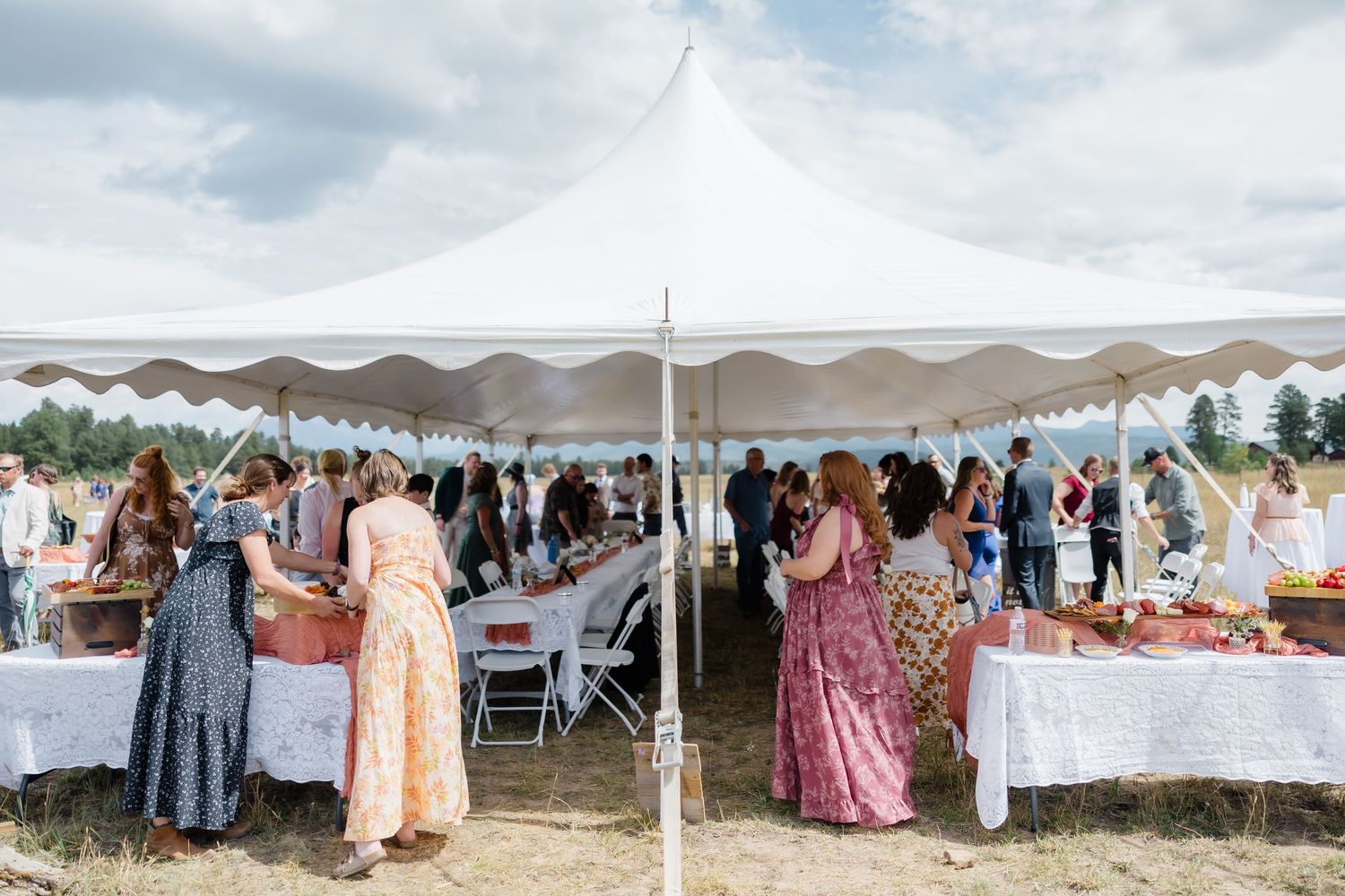 Guests gather under a white tent at an outdoor catered event, with long tables and food displays on a sunny day.