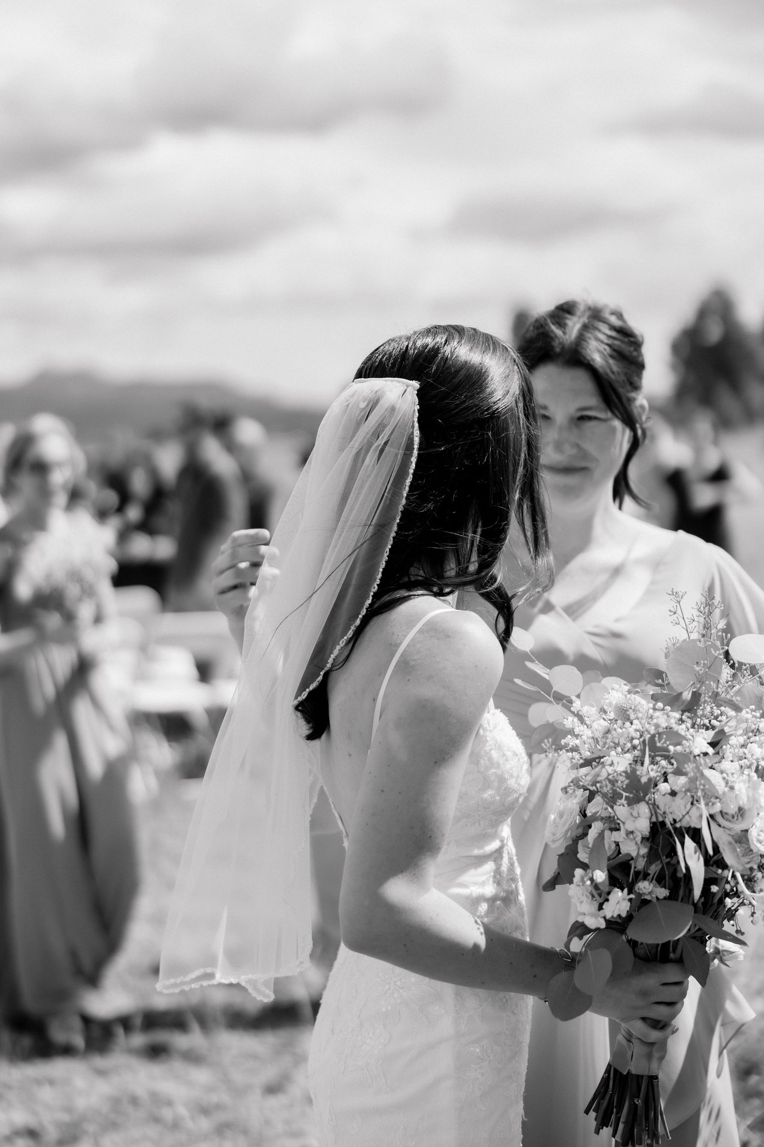 Black and white photo of two brides sharing a tender moment, one wearing a veil and holding a wildflower bouquet at an outdoor wedding.