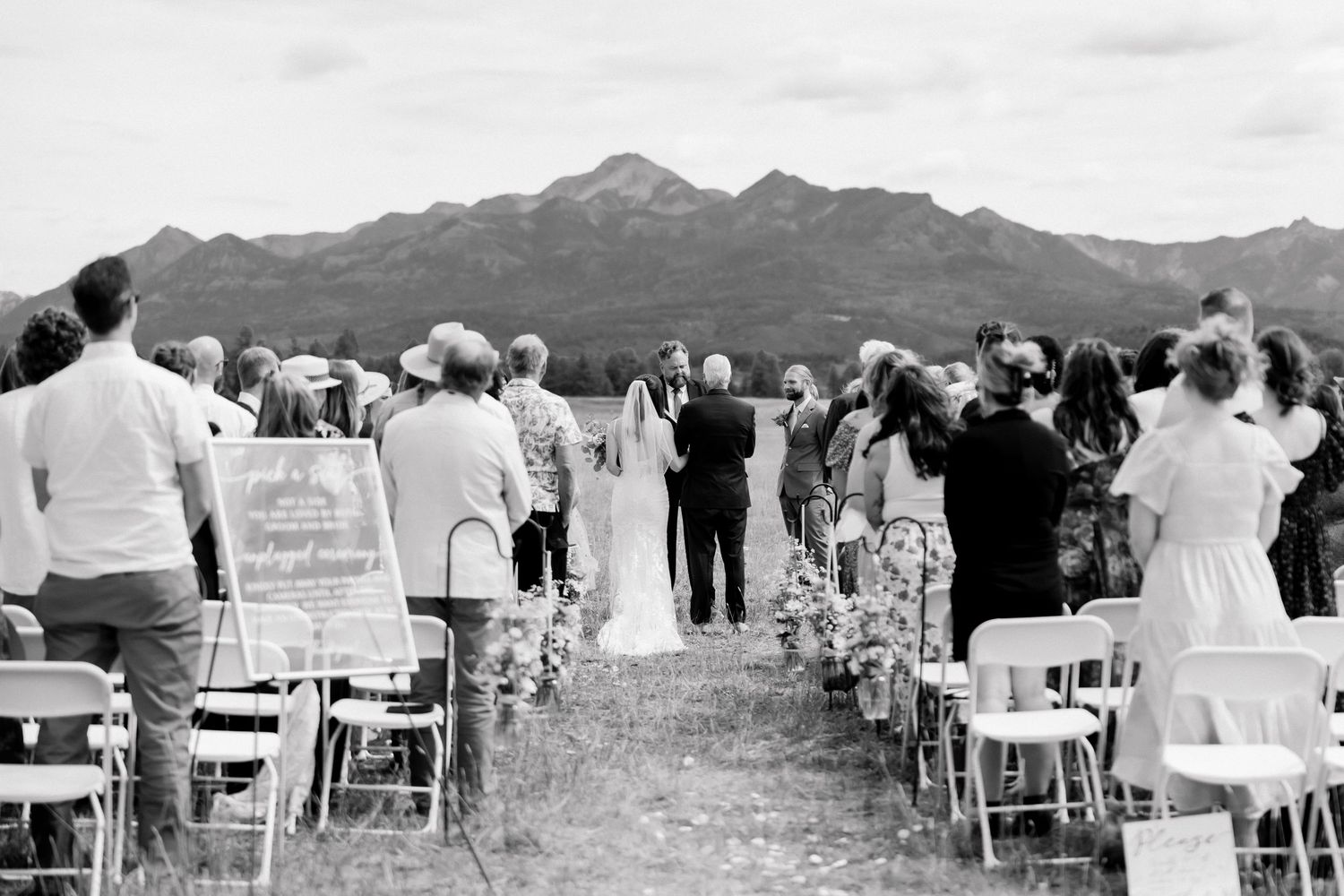 Outdoor wedding ceremony with guests seated in white chairs, couple at altar, and stunning mountain backdrop in black and white.