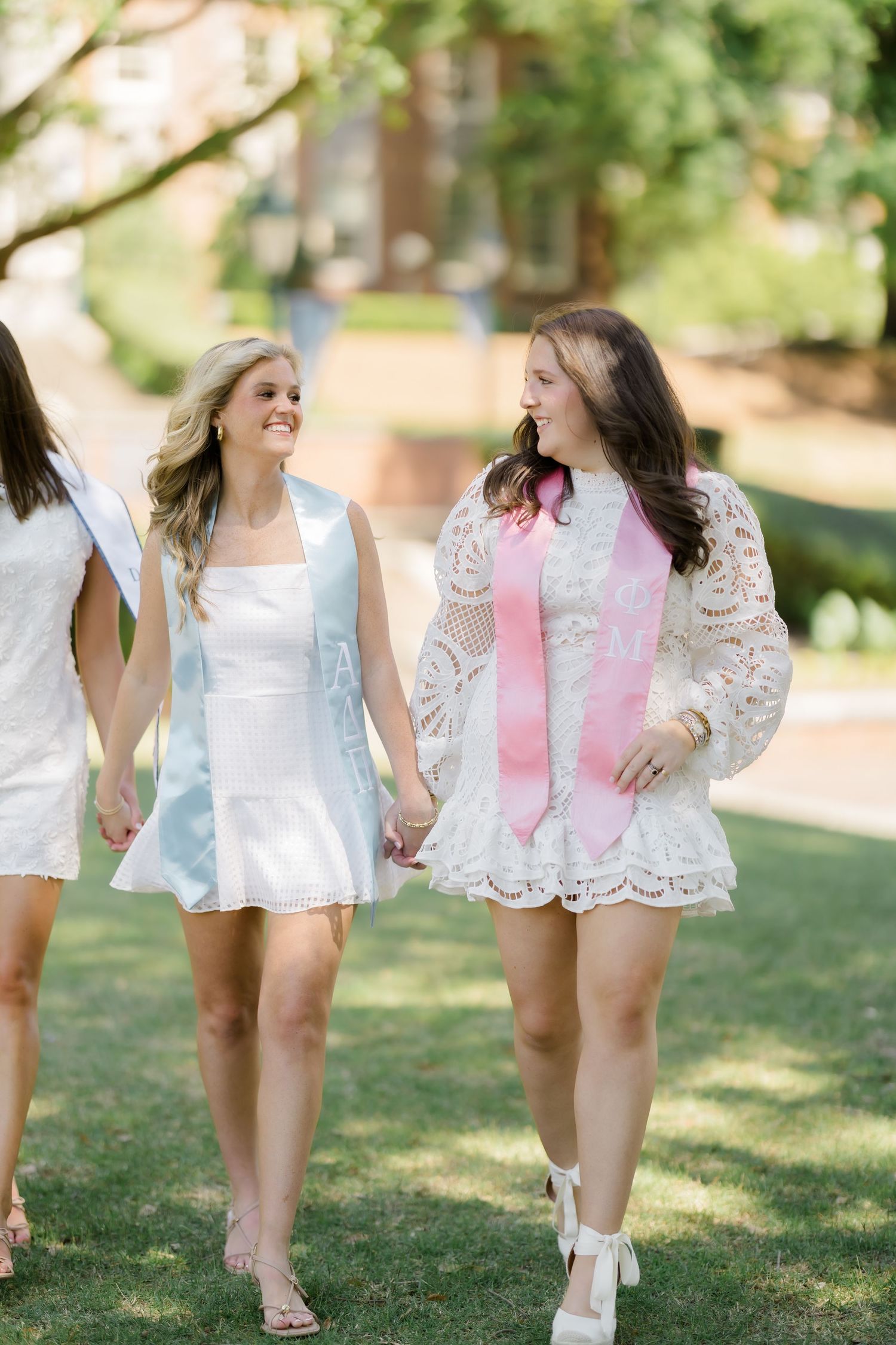Two young women in white dresses holding hands and smiling, one wearing a pink graduation sash, walking outdoors.