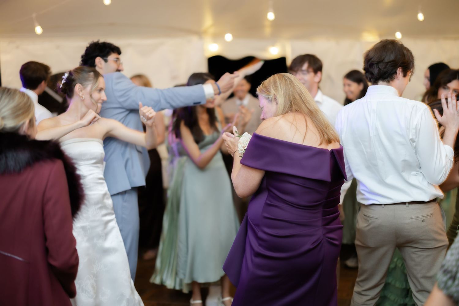 Guests dancing joyfully at a wedding reception under a white tent, including bride in white gown and woman in purple dress.