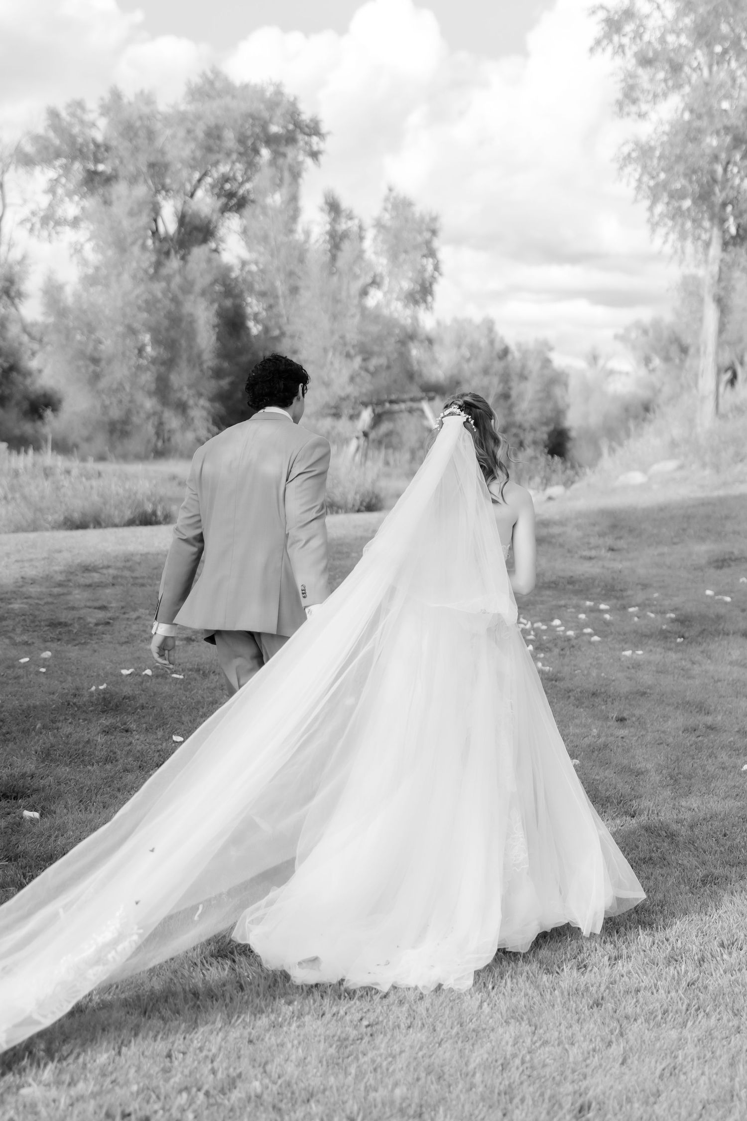 Bride and groom walking hand in hand across green lawn, cathedral veil trailing behind.