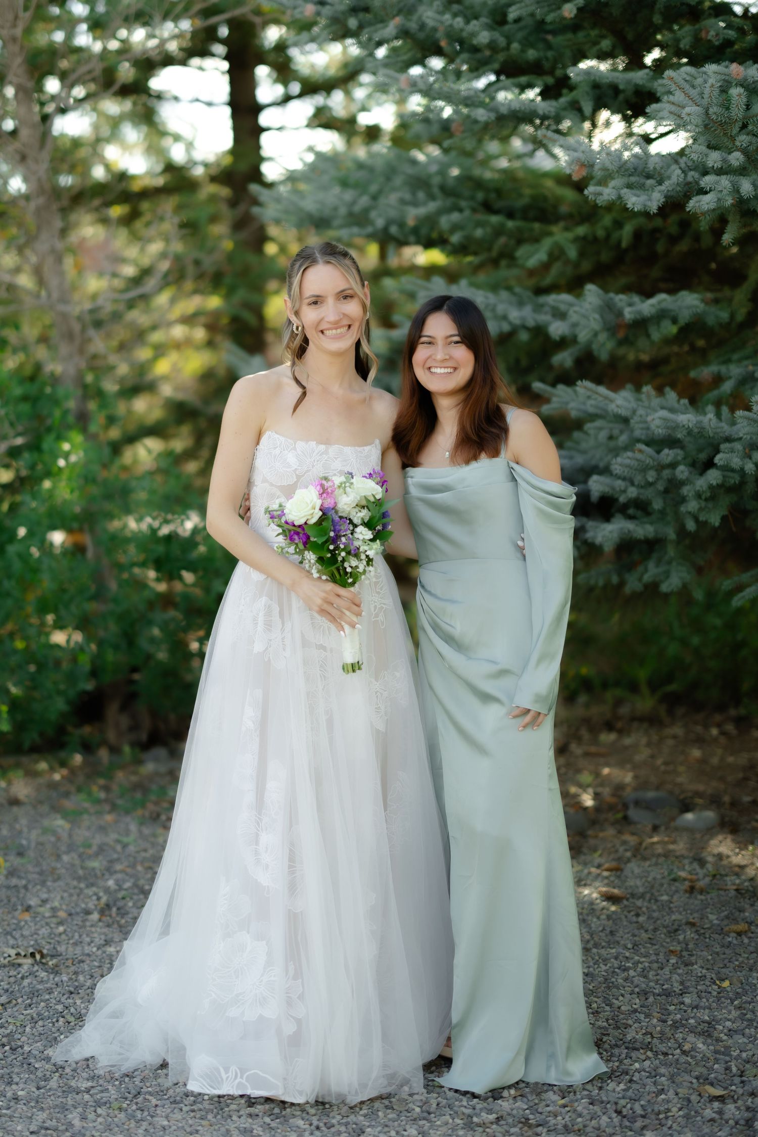 Bride in white lace gown holding floral bouquet poses with bridesmaid in sage green dress outdoors.