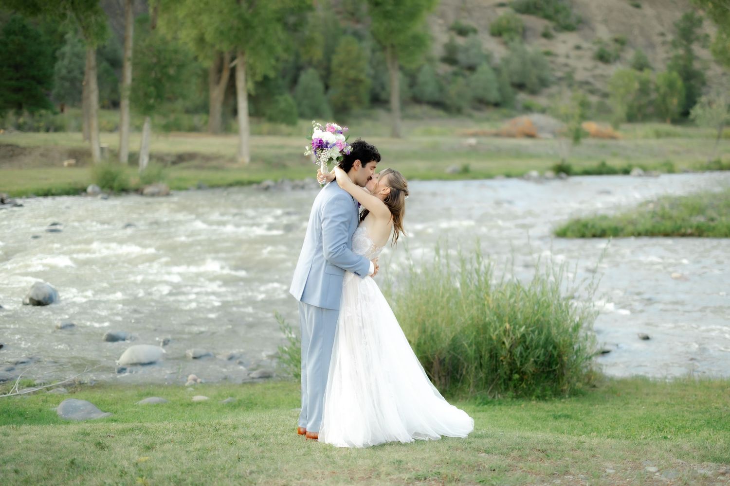 Bride and groom sharing a romantic kiss by a mountain river, bride holding a colorful bouquet in a white gown.
