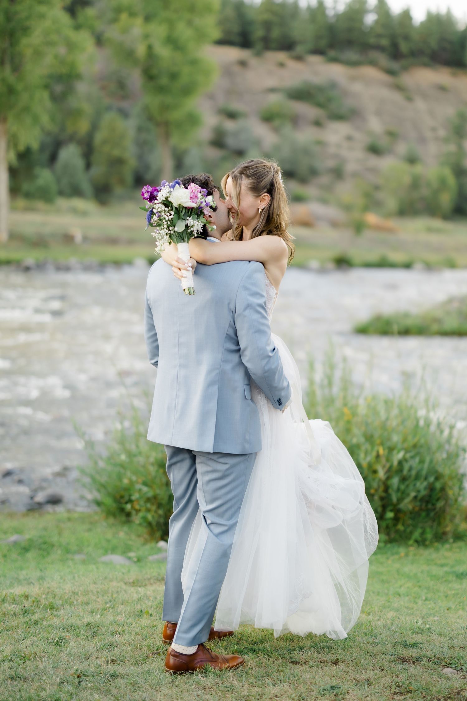 Bride and groom embracing outdoors near a river, she holds a purple bouquet in a white gown, he wears a light gray suit.