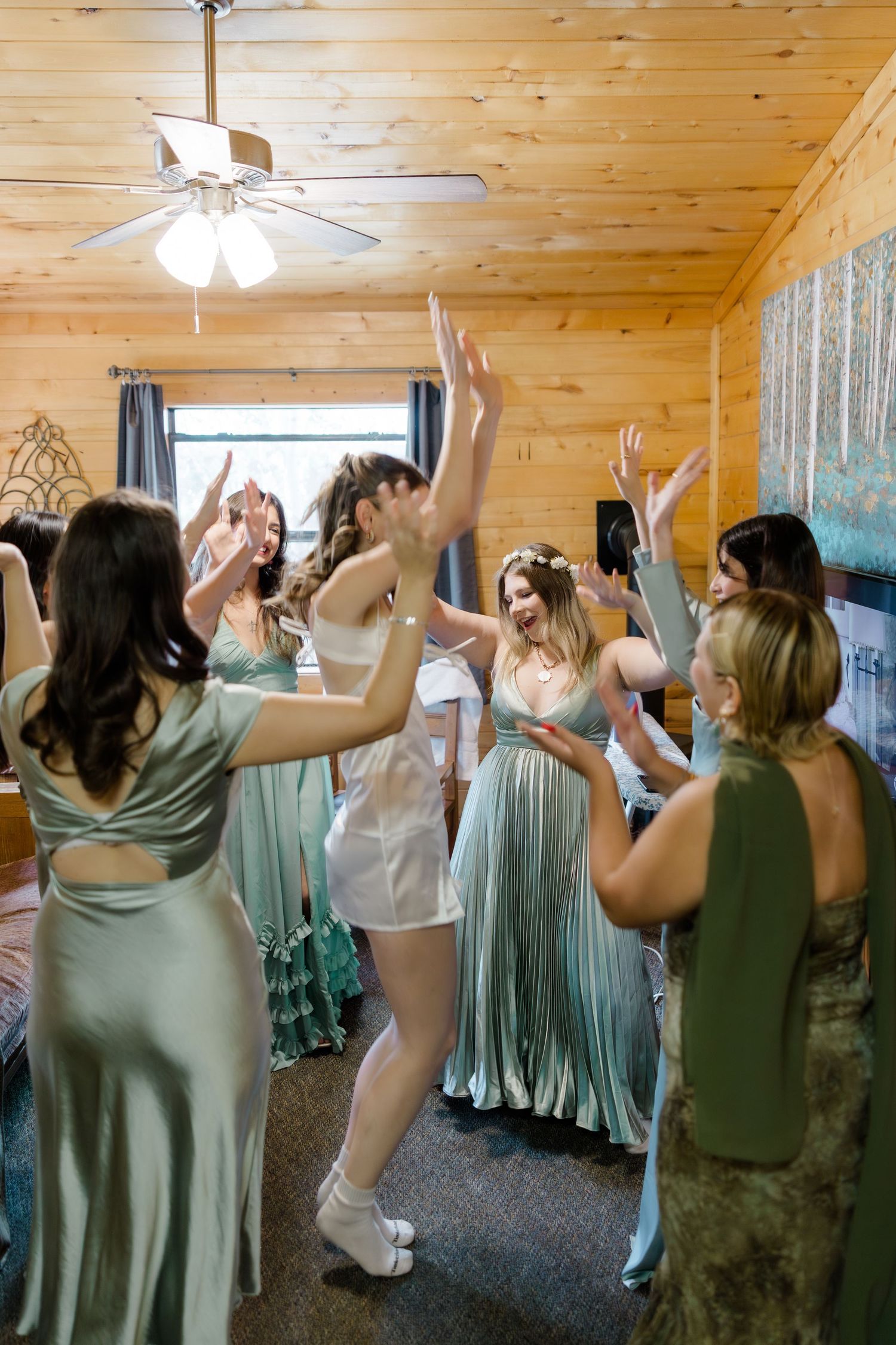 Bride in white dancing with bridesmaids in sage green dresses inside a rustic wooden cabin before the wedding.