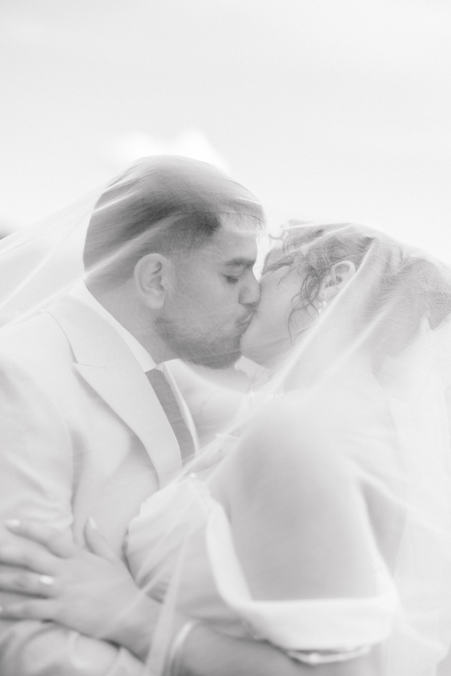 Black and white photo of a bride and groom sharing a romantic kiss under a flowing wedding veil.