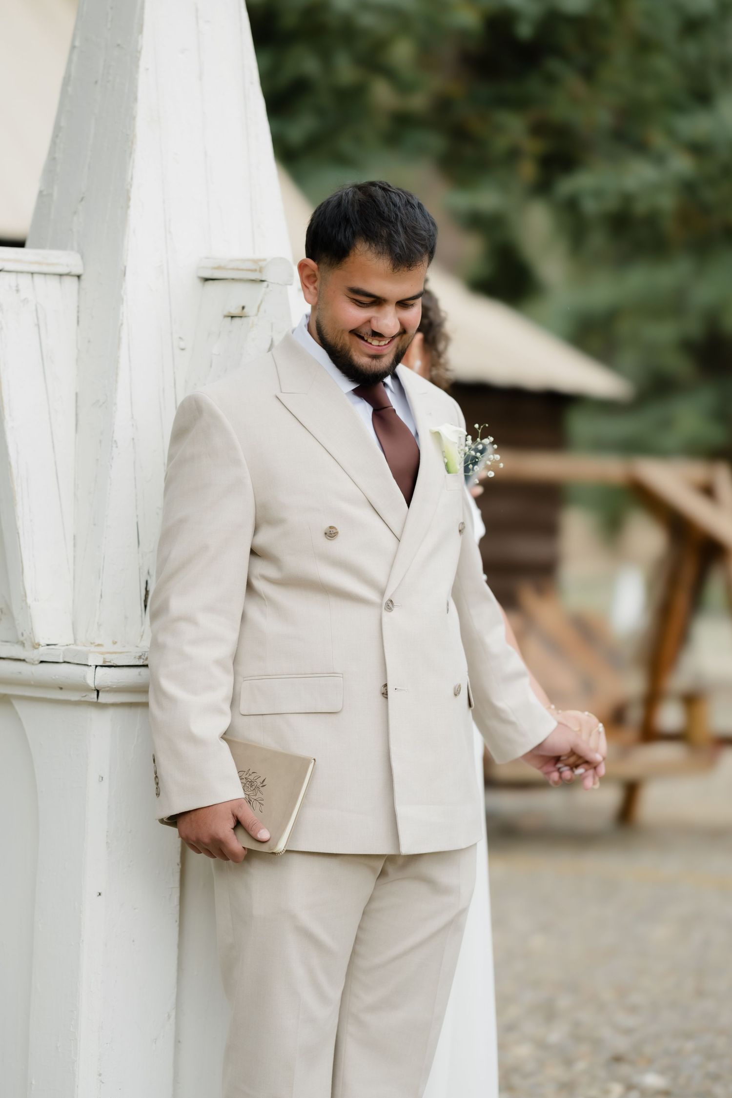 Smiling groom in beige double-breasted suit with boutonniere holding a book outdoors at a wedding venue.