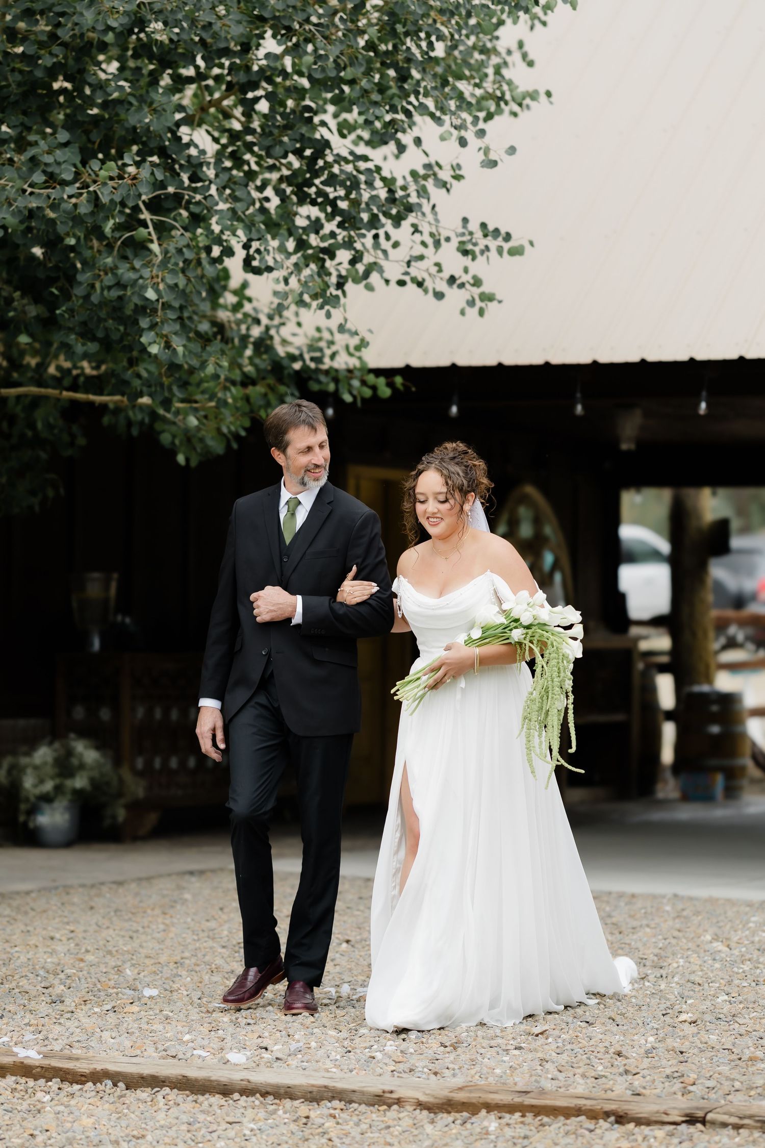 Bride in white off-shoulder gown holds bouquet while walking with groom in black suit on gravel path.