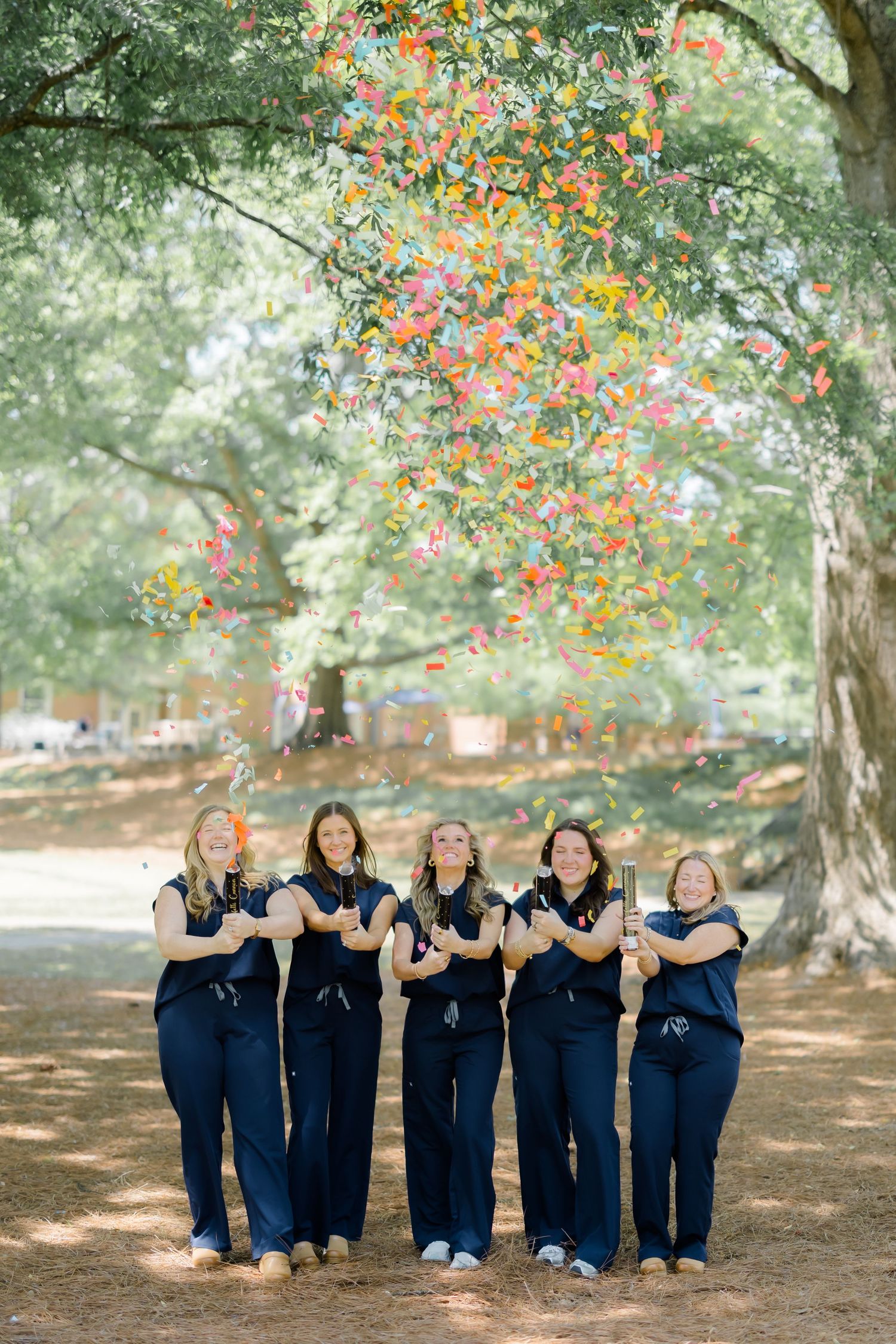 Six women in matching navy jumpsuits laugh as colorful confetti falls around them outdoors beneath large shade trees.