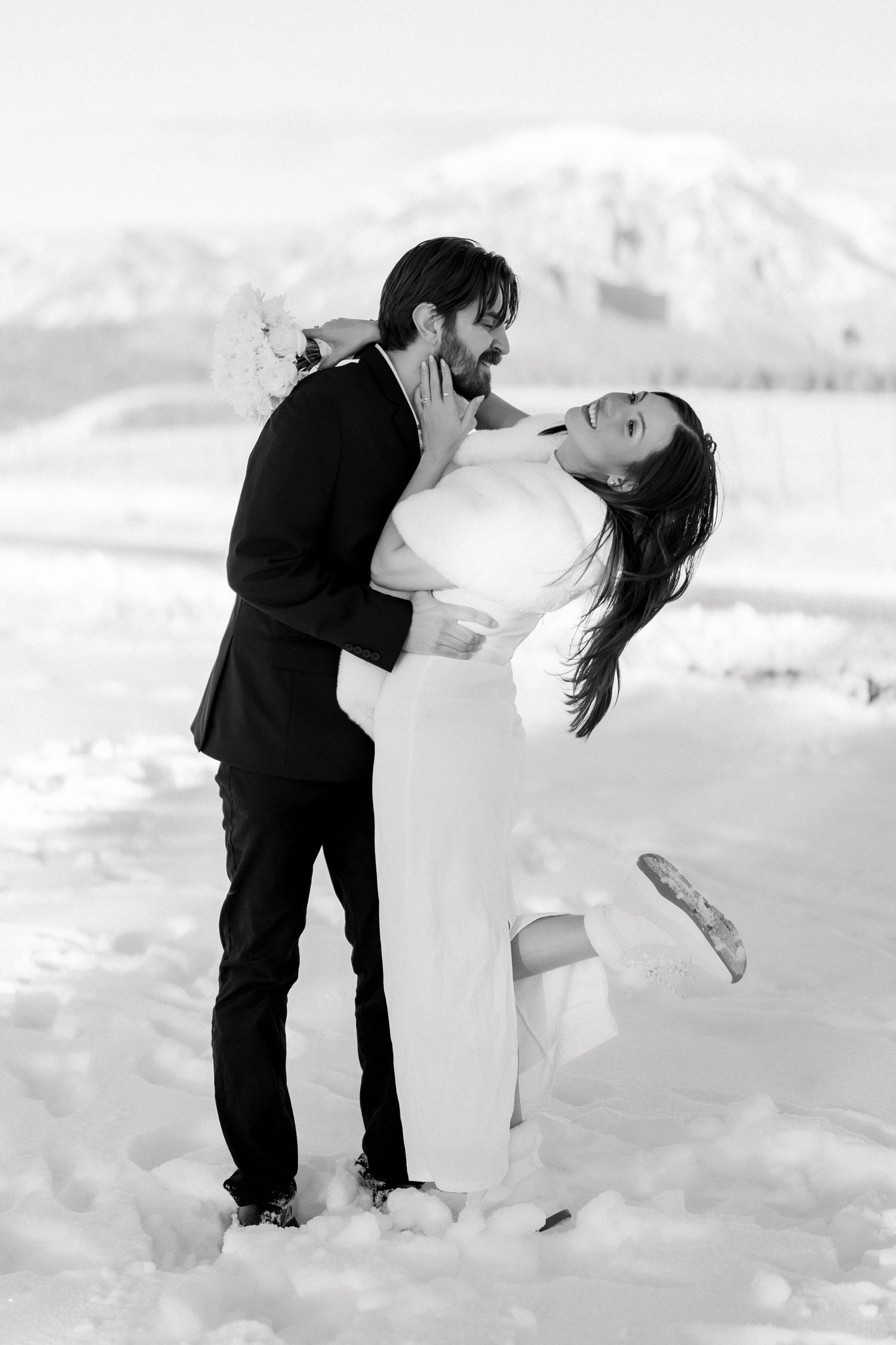 Black and white photo of groom dipping bride dramatically in snowy open mountain landscape.