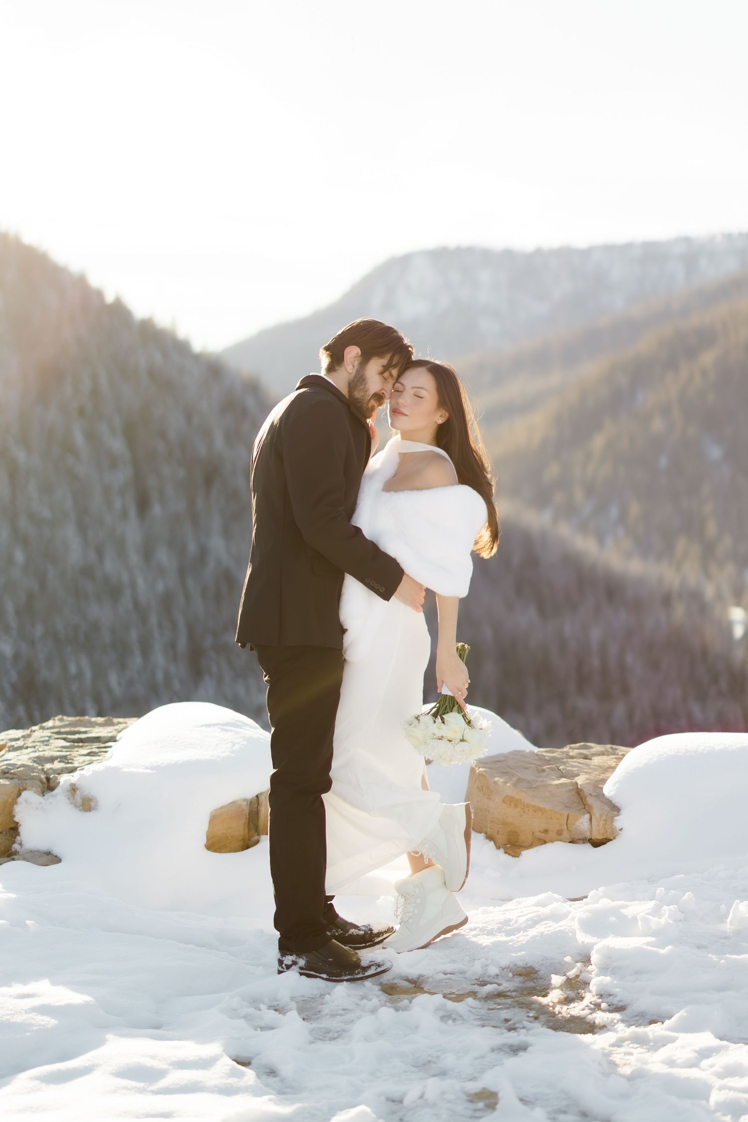 Bride and groom embrace on snowy mountain overlook, bride in white gown with bouquet, groom in black suit, sunny winter backdrop.
