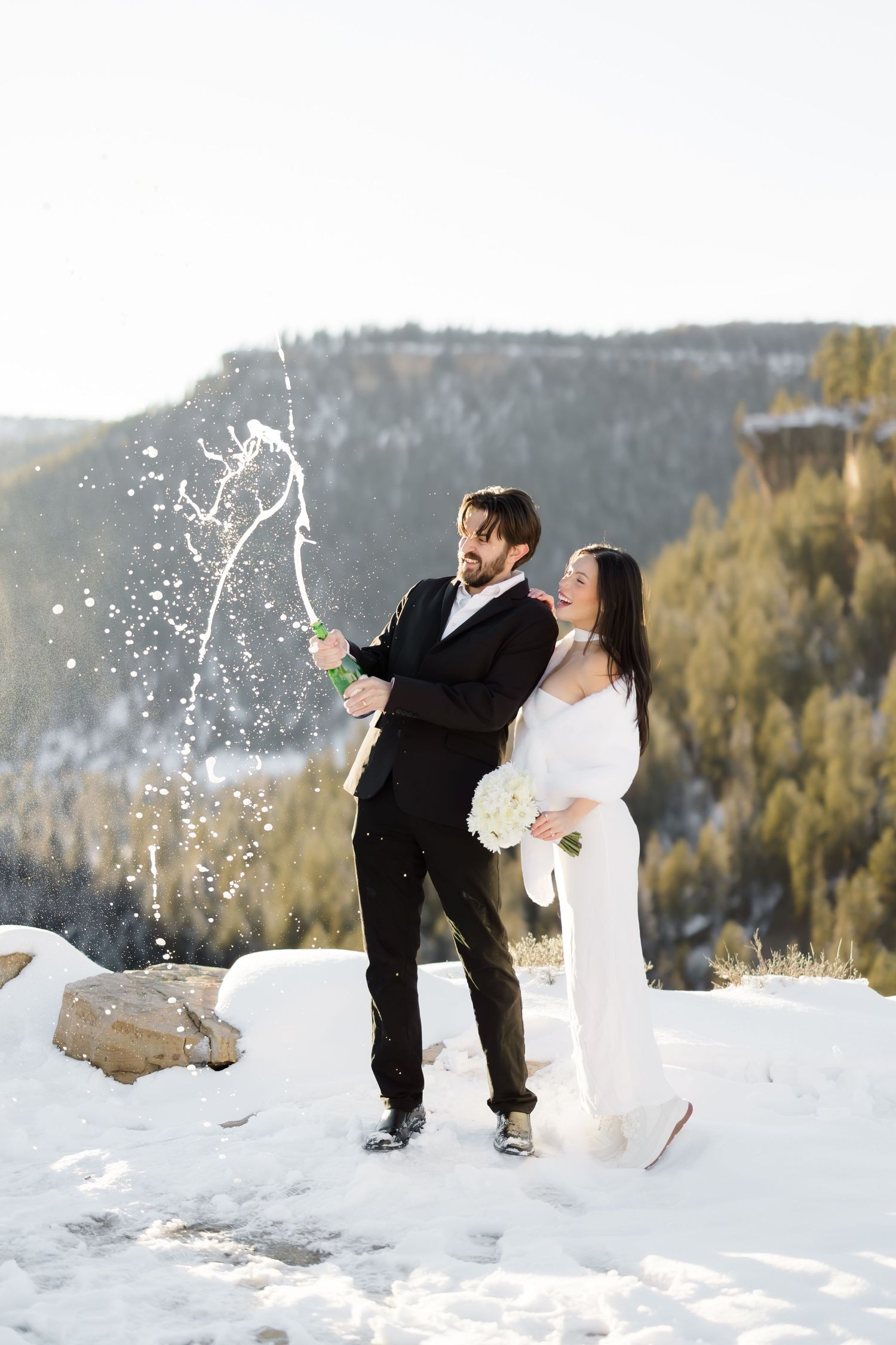 Bride and groom popping champagne in snowy mountain setting, laughing joyfully in winter wedding attire.