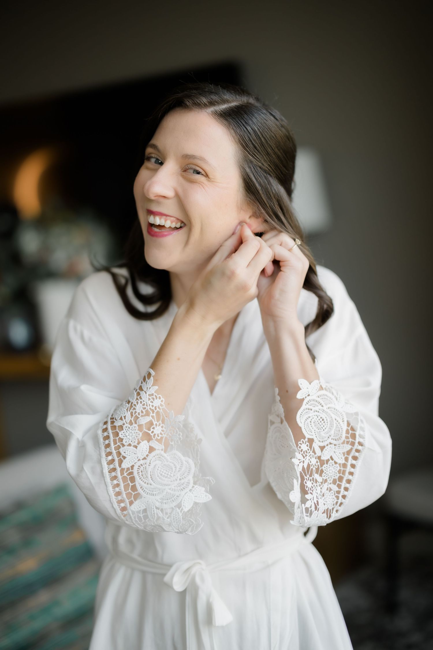Smiling bride in white lace robe putting on earrings while getting ready for her wedding day.