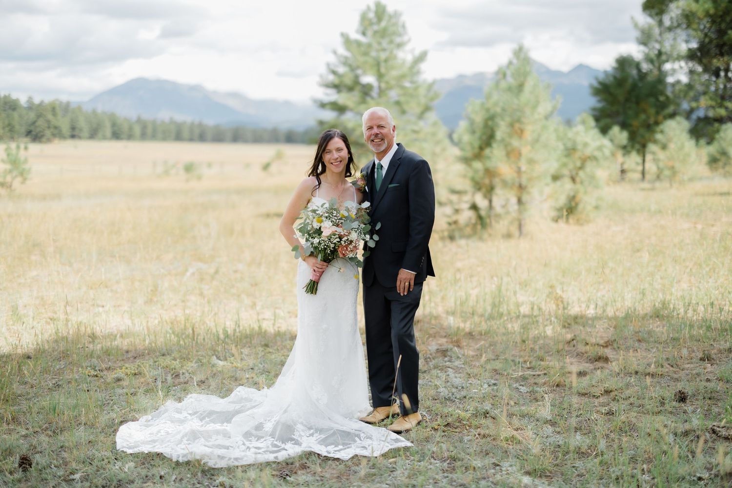 Bride in lace gown with bouquet stands with groom in navy suit in a golden meadow with mountain views.