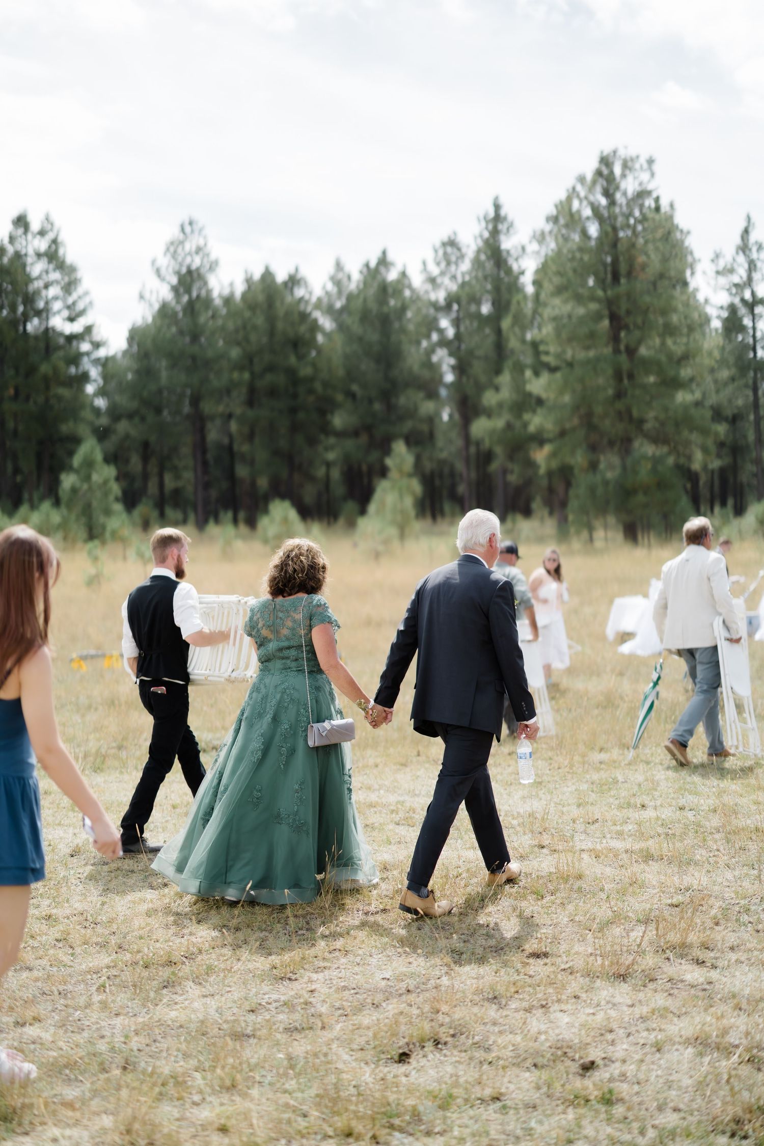 Wedding guests walk hand-in-hand across a rustic field, with a woman in a green gown and man in a suit leading the procession.