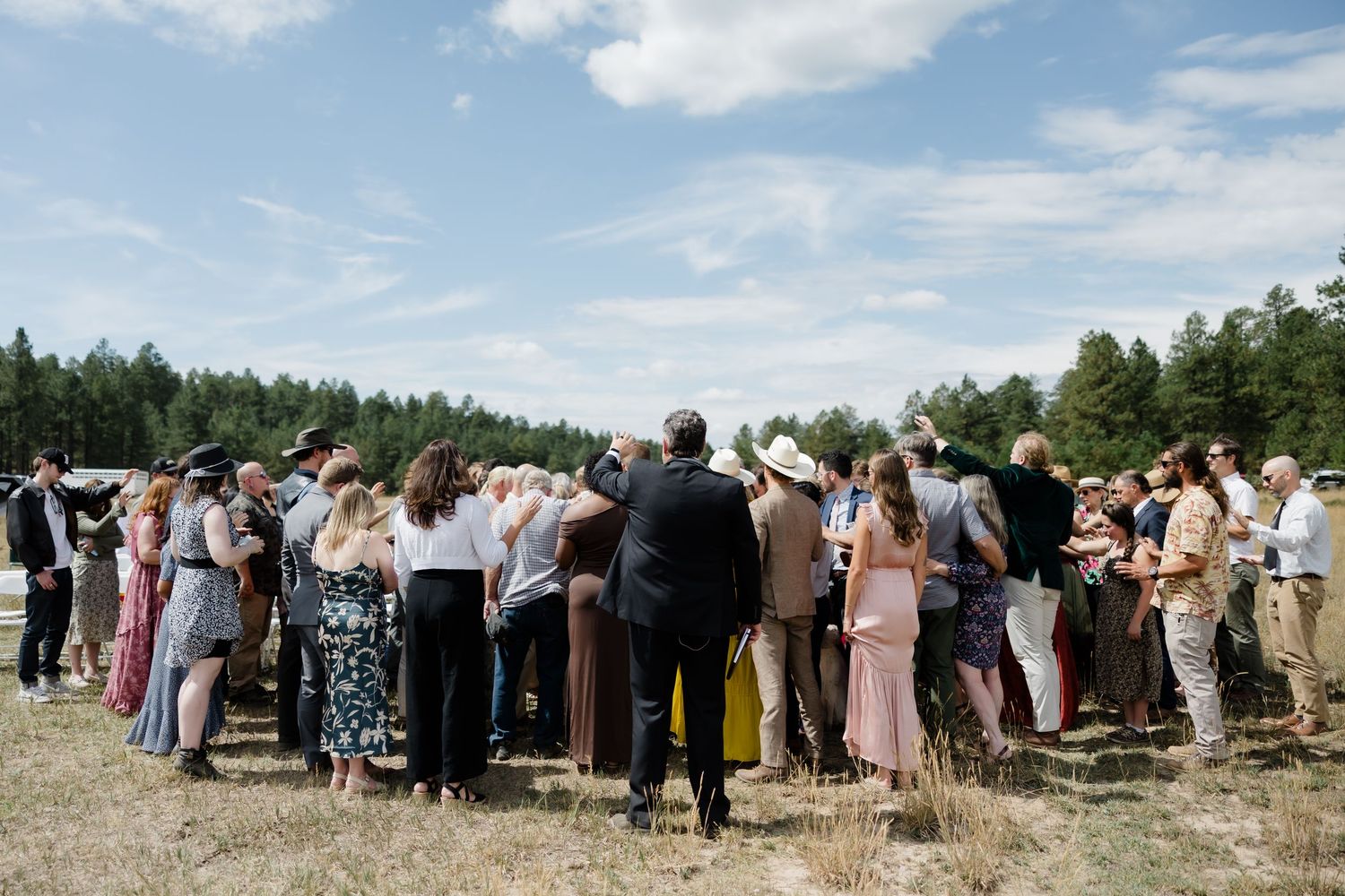 Outdoor wedding guests gathered in a field surrounded by pine trees under a partly cloudy blue sky.