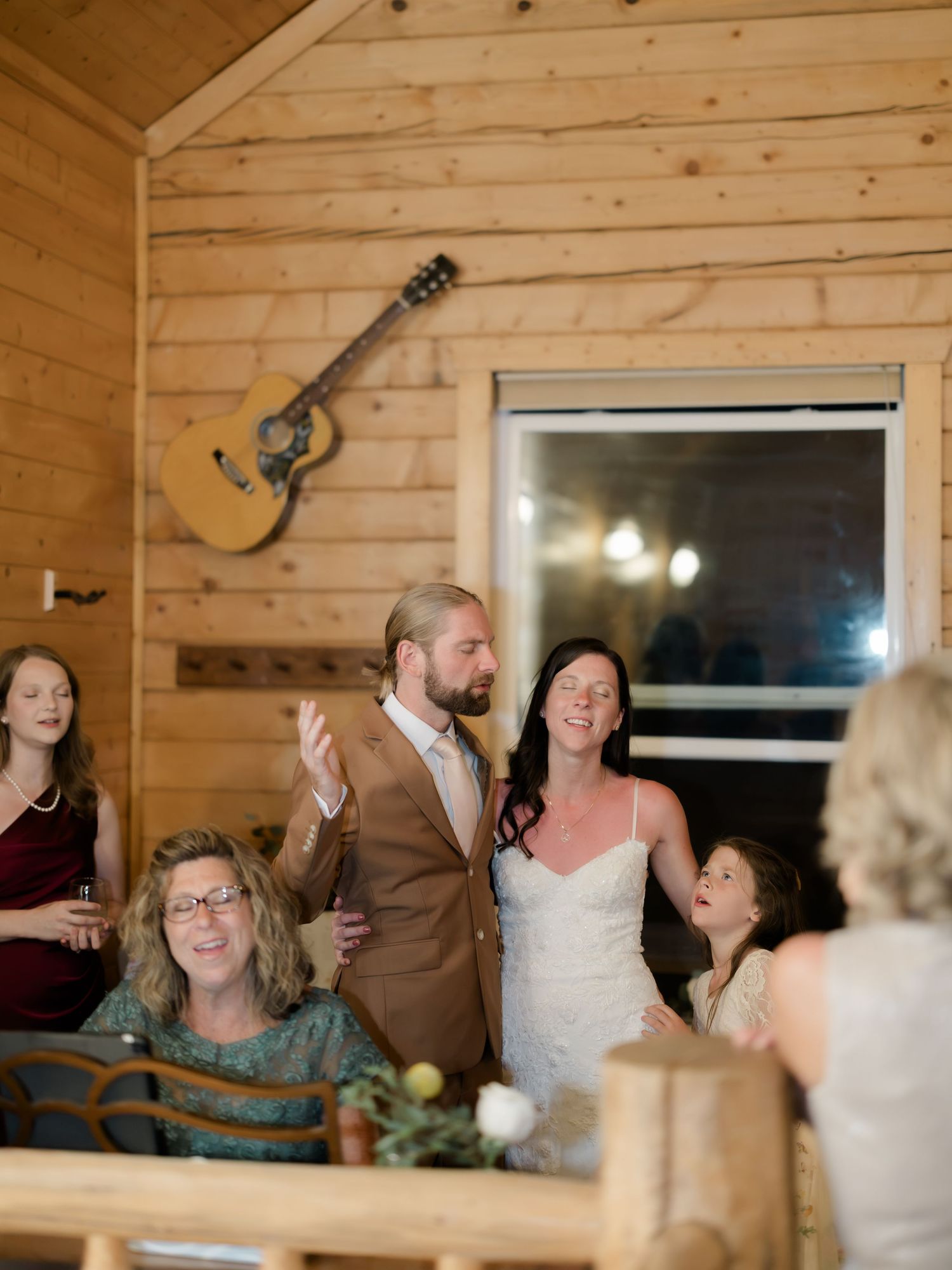 Couple in wedding attire smiling with family inside a rustic wood cabin venue with guitar on wall.