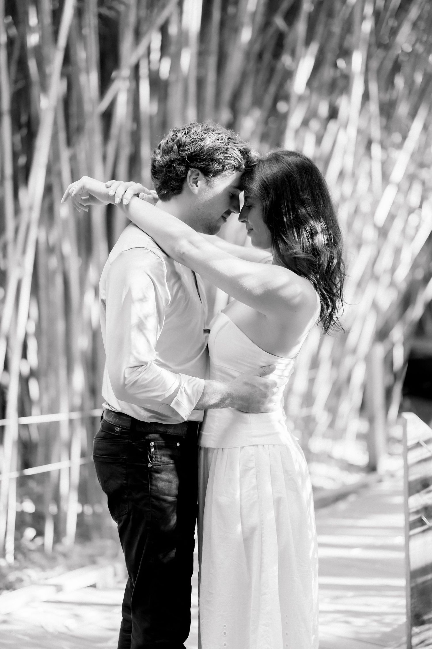 Black and white photo of a romantic couple embracing and almost kissing among bamboo trees outdoors.