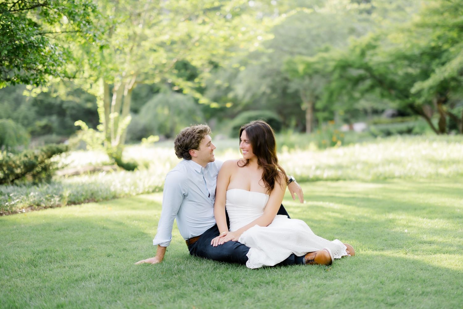 A couple sits together on lush green grass in a garden, gazing lovingly at each other on a sunny day.