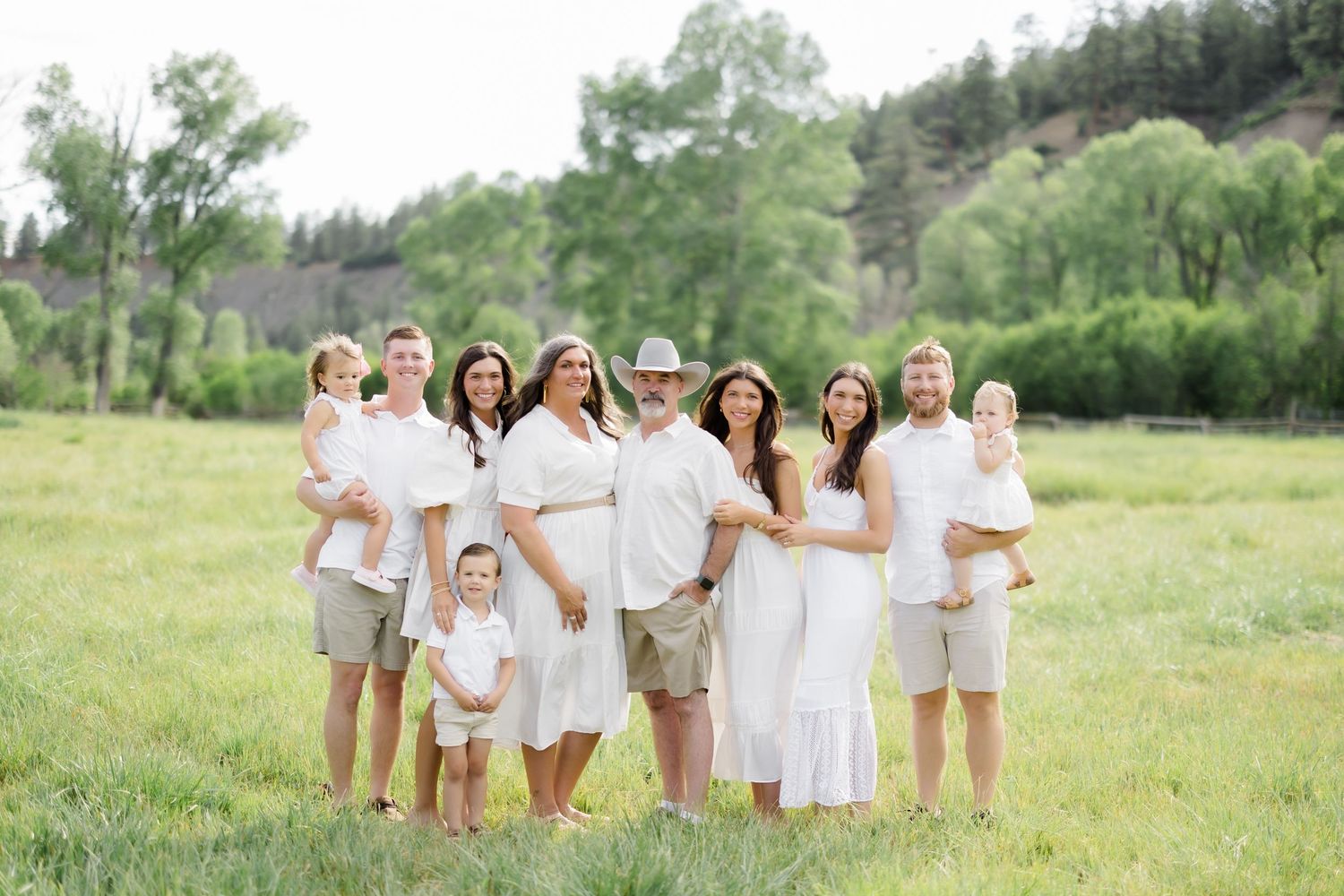 Large family posing outdoors in white and tan outfits in a lush green meadow with trees in the background.