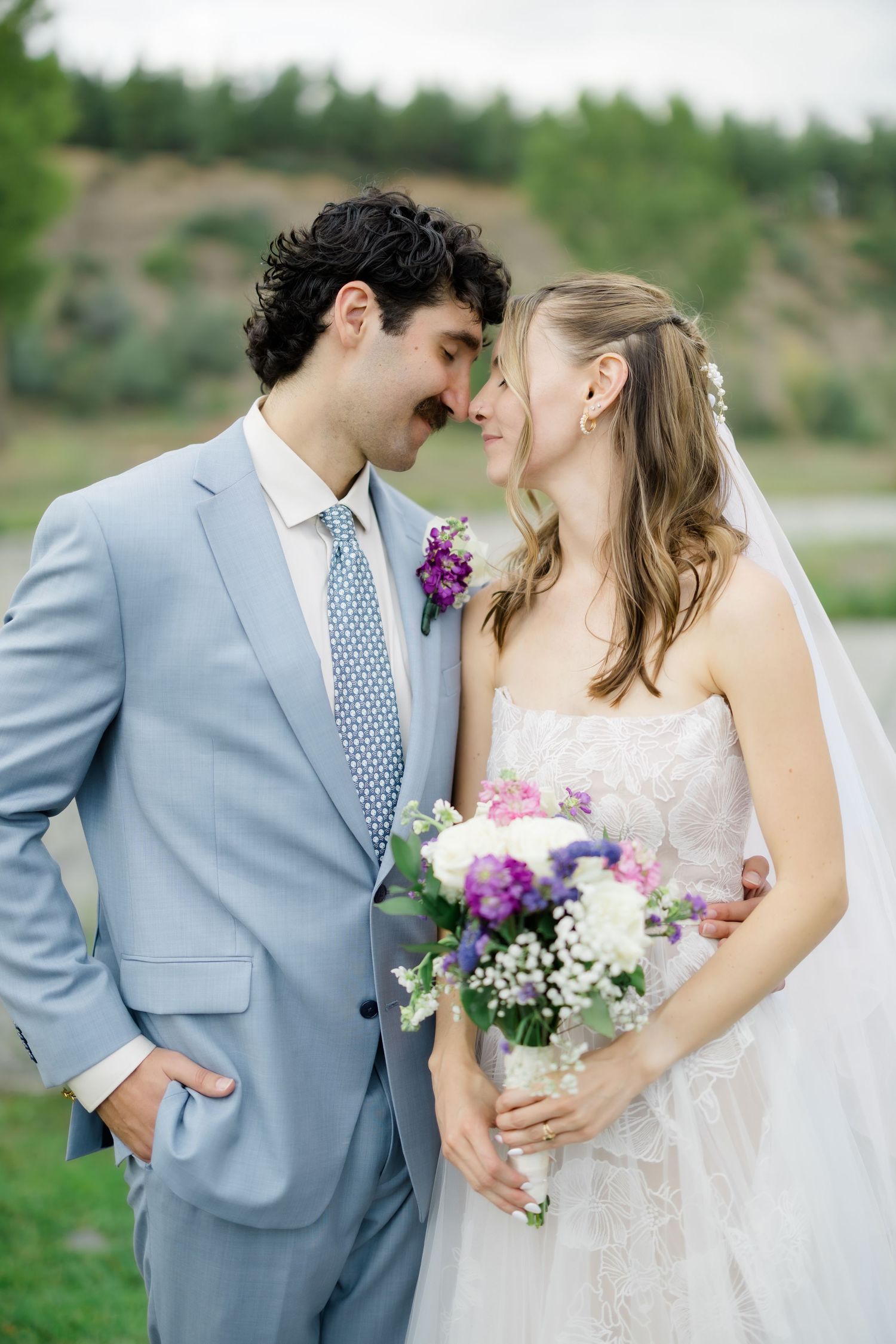 Bride and groom share a romantic moment, touching foreheads outdoors, bride holding purple and white bouquet.
