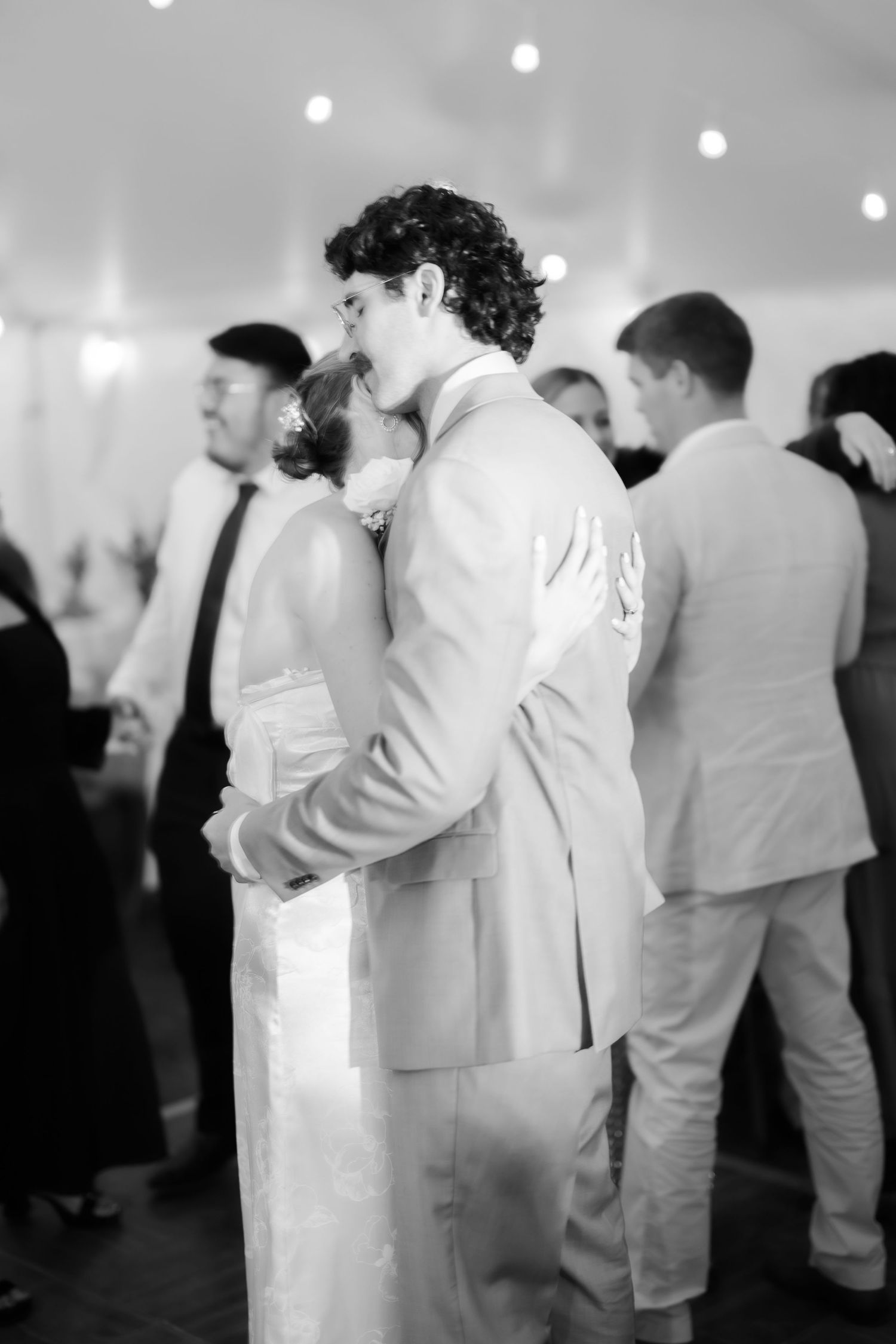 Black and white photo of bride and groom sharing a kiss on the dance floor surrounded by wedding guests.